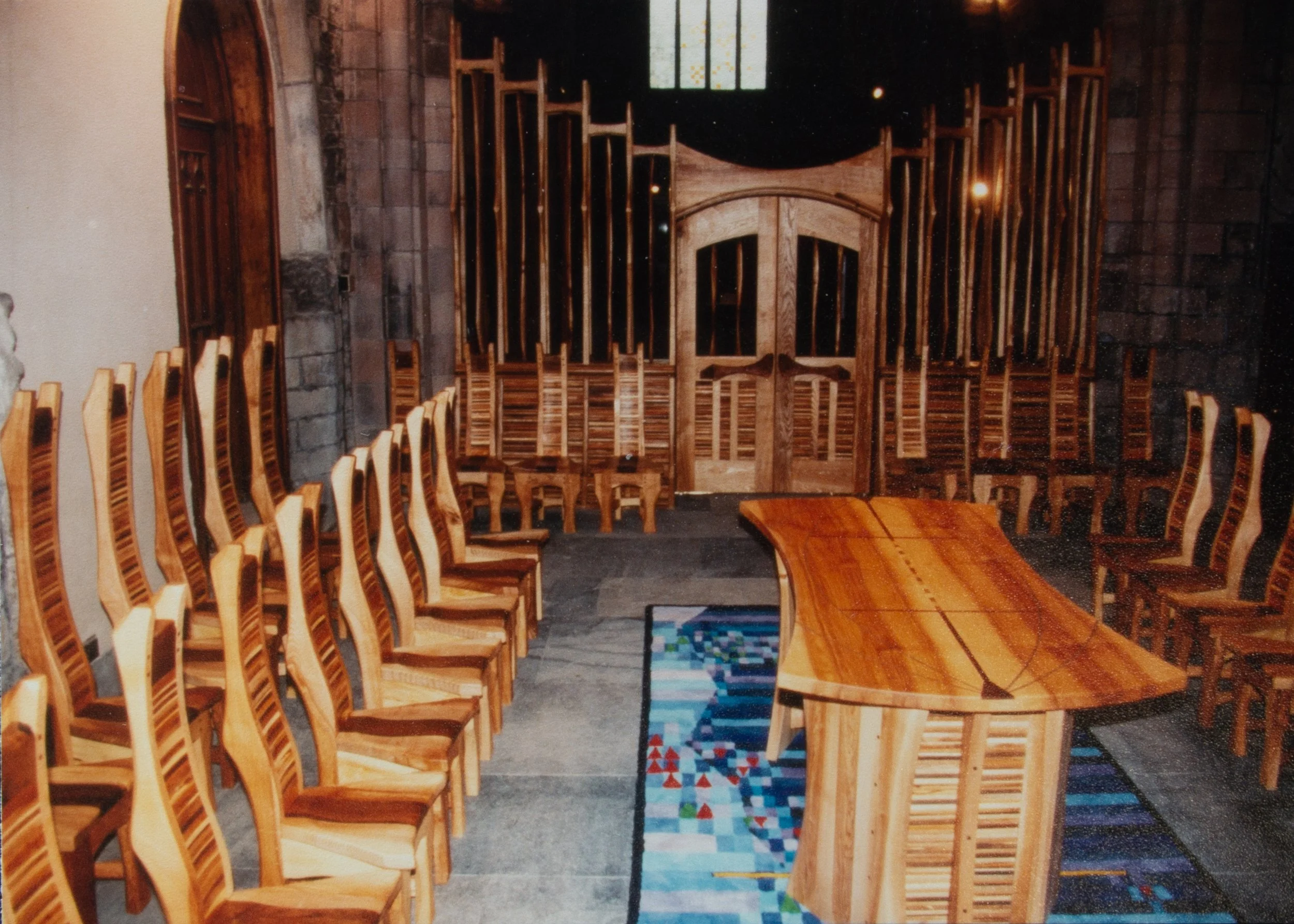 A room with wooden chairs arranged in a U-shape around a large, curved wooden table, with an ornately carved wooden door and stone walls in the background.