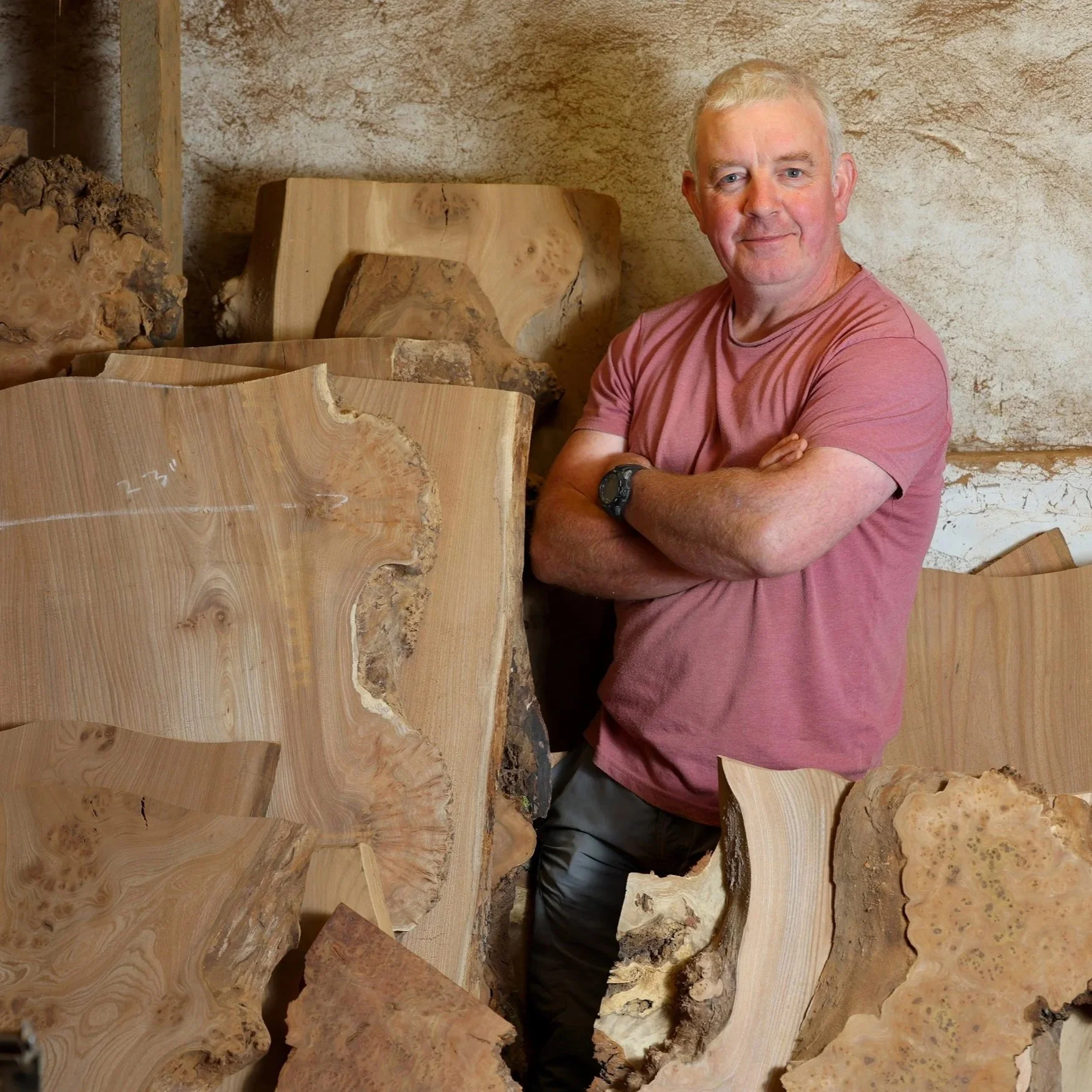 A man in a pink t-shirt with crossed arms standing in front of walnut wood slabs in a workshop.