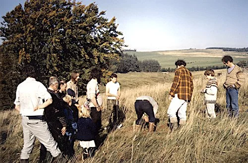 A group of people outdoors in a grassy field, some looking at the ground and others standing, with rolling hills and trees in the background.