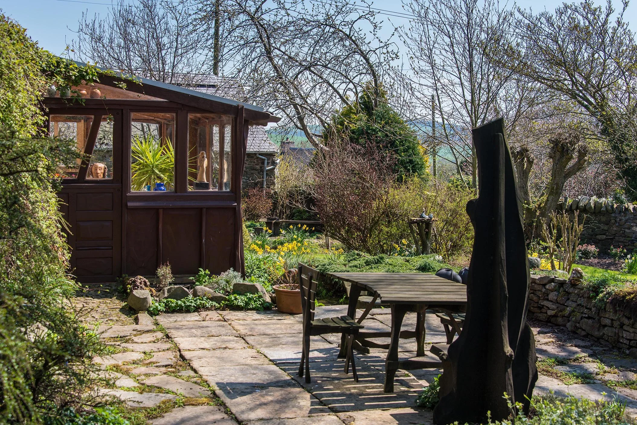 A garden with a wooden shed, stone pathway, outdoor table with chairs, trees, and bushes in springtime.