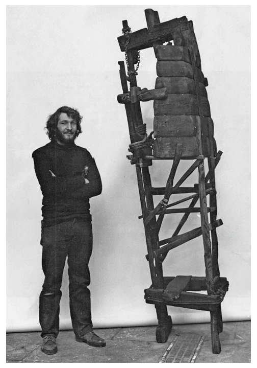 A man with curly hair and a beard standing next to a tall, oddly constructed wooden chair with a large wooden seat or backrest, set against a plain white background.