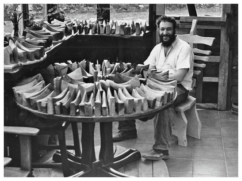 A man with a beard sitting at a round table with numerous wooden shoe lasts, inside a workshop with windows.