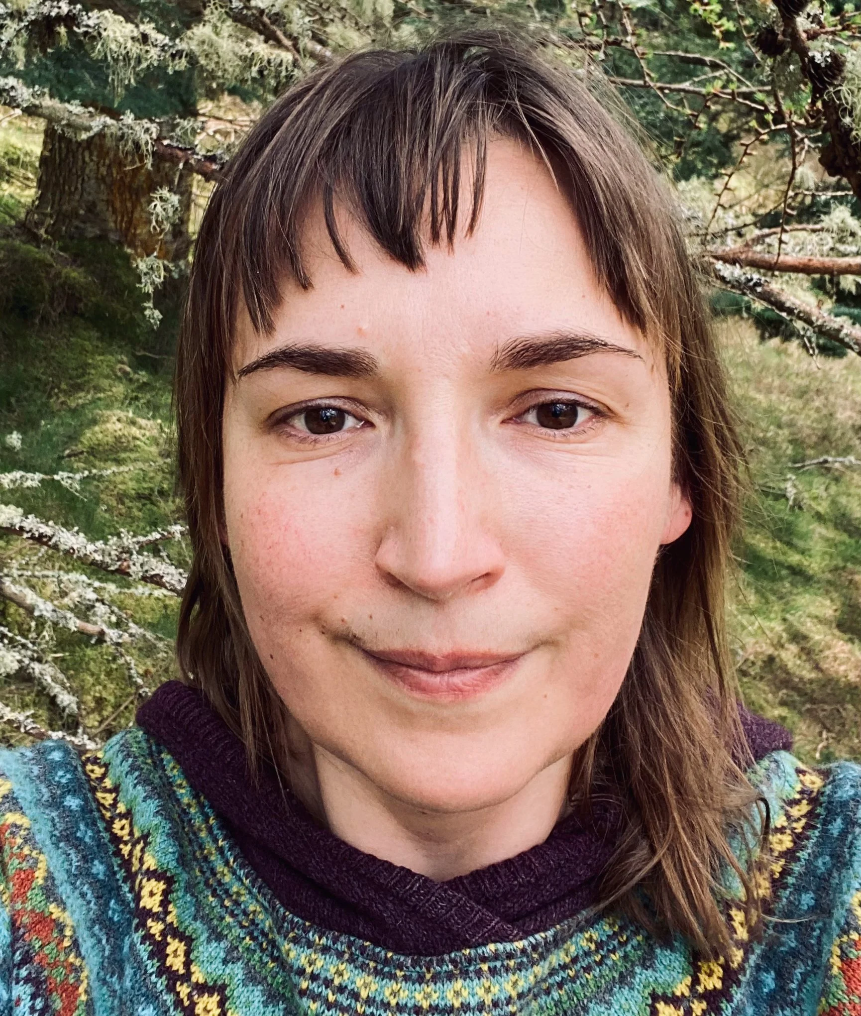 Close-up of a woman with brown hair and brown eyes, wearing a colorful, patterned sweater, outdoors among trees and greenery.