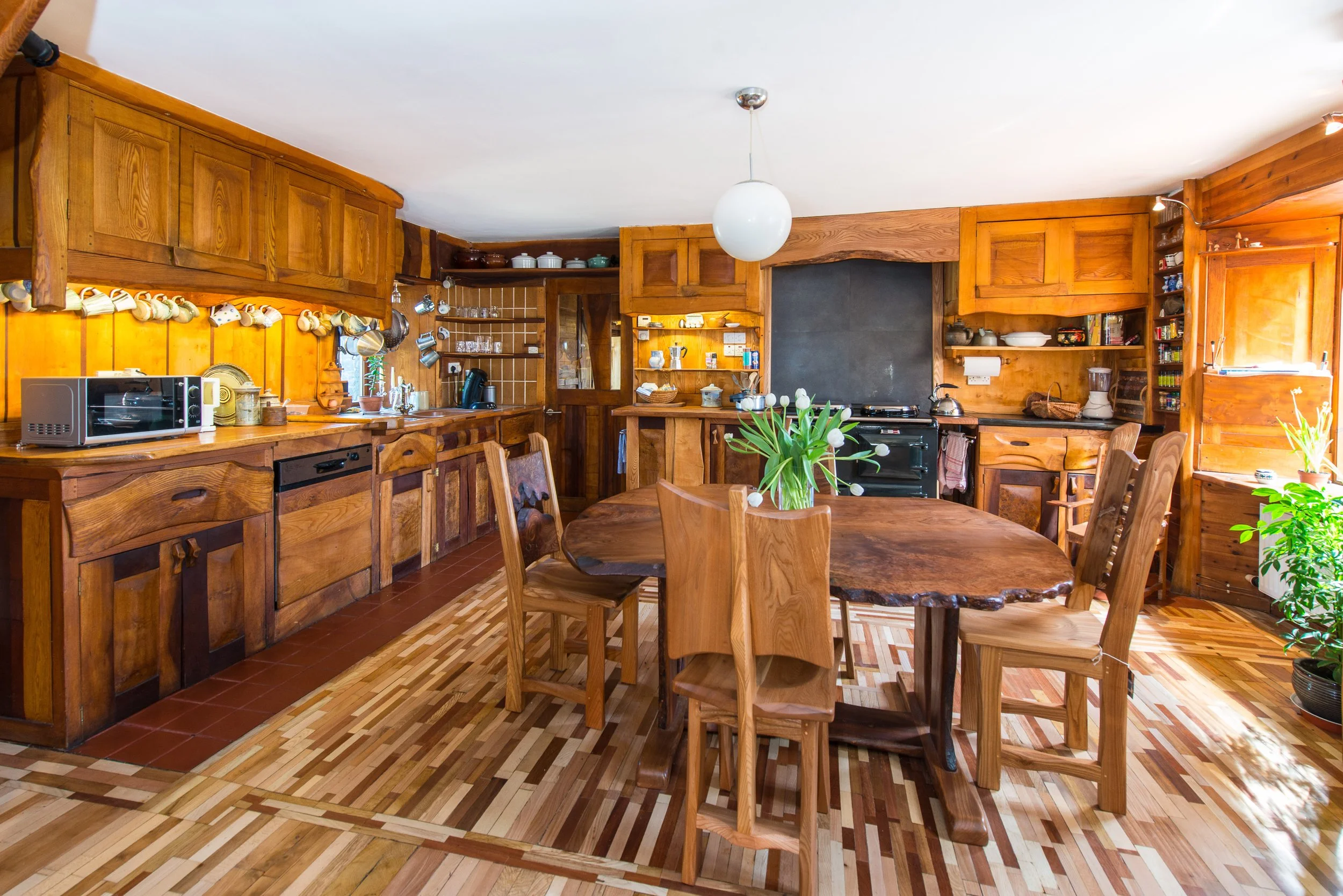 Wooden kitchen and dining area with a round wooden table, six matching chairs, a bouquet of white tulips on the table, open shelves with dishes, a black stove, and a window with plants.