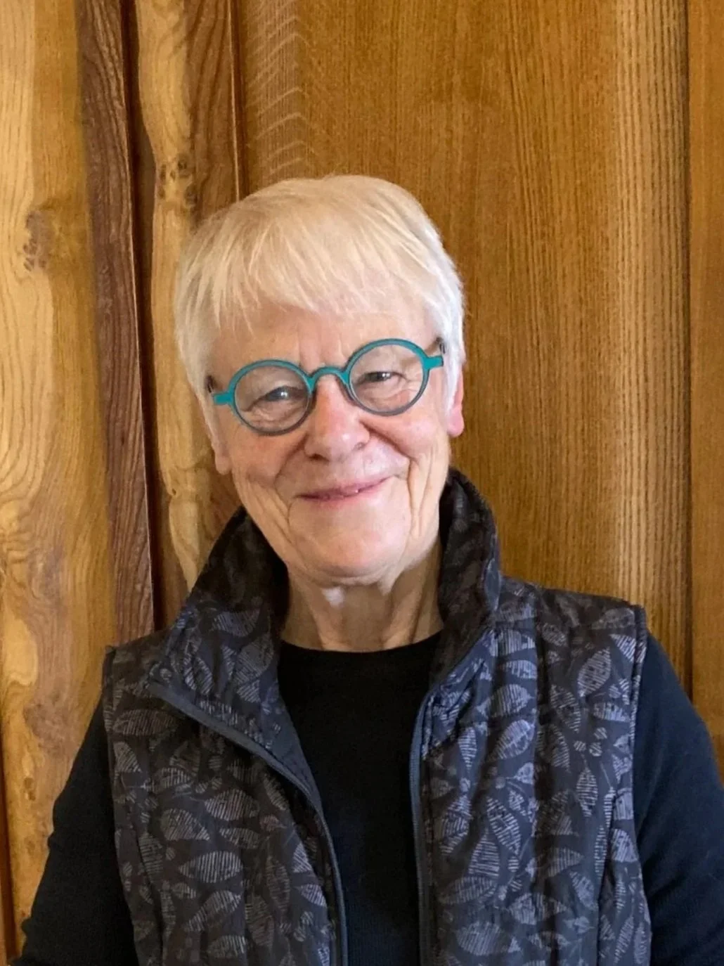 An elderly woman with short white hair and round teal glasses, smiling in front of a wood-paneled wall, wearing a black shirt and a patterned black vest.