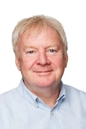Headshot of a middle-aged man with light hair, wearing a light blue collared shirt, smiling against a plain white background.