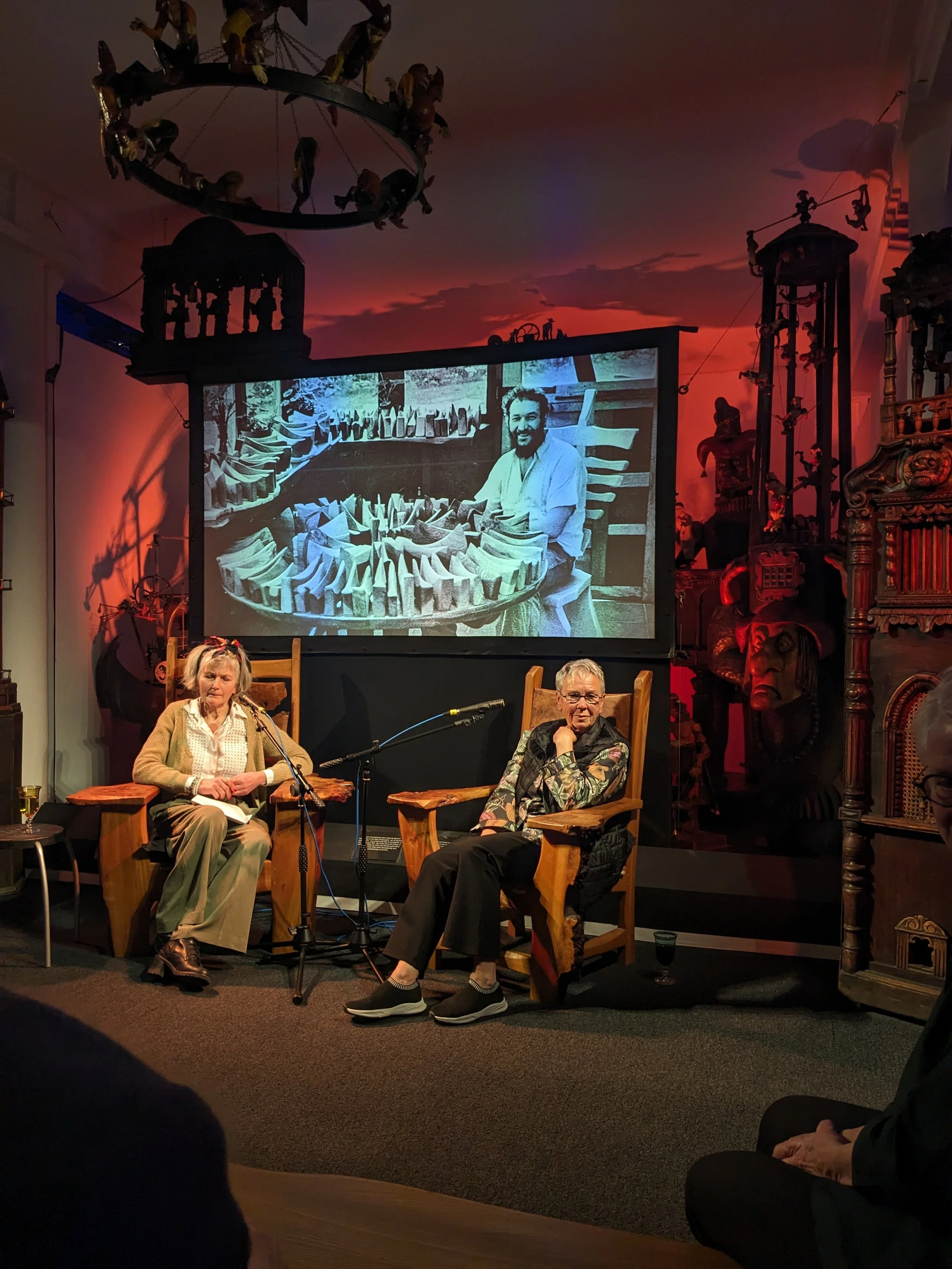 Two people are seated on stage in front of a large screen showing a black-and-white photo of a man with a collection of objects around him. The setting appears to be a talk or presentation with microphones, chairs, and a decorative background with African art and masks.