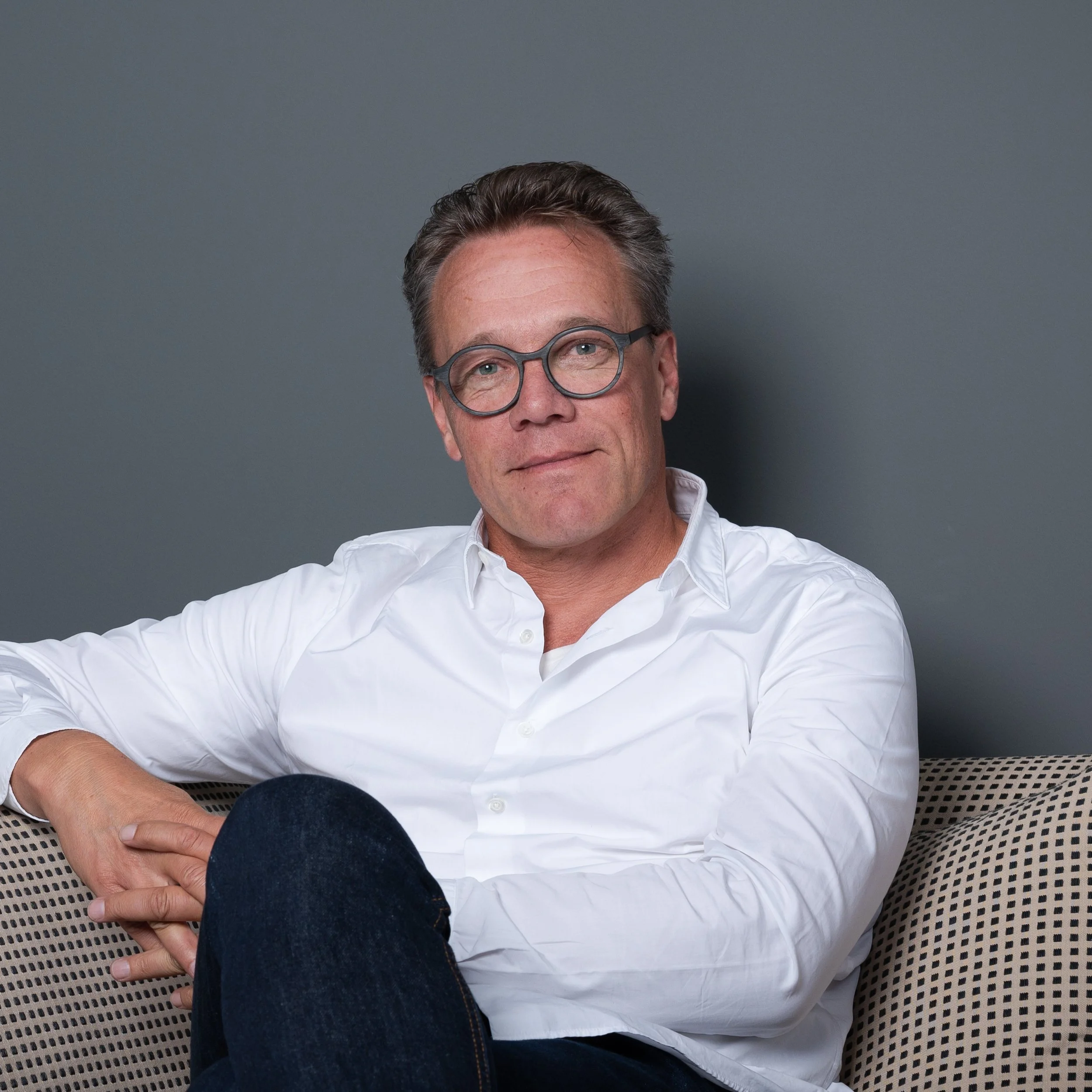 Ein Mann mit Brille, weißem Hemd und dunkler Hose sitzt entspannt auf einem Stuhl vor einer grauen Wand.