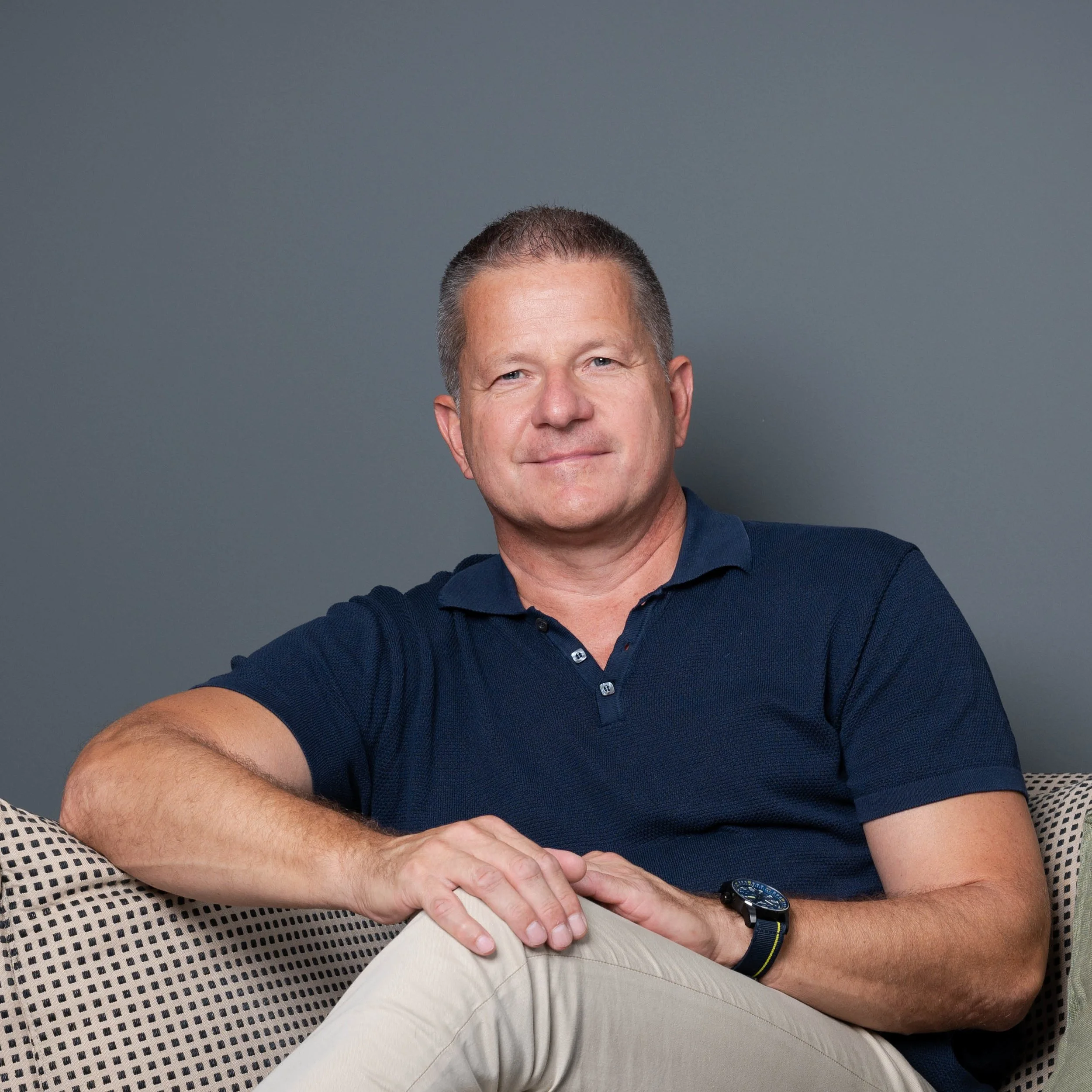 Mann sitzt auf einem Sofa vor einer grauen Wand, trägt ein dunkelblaues Polo-Shirt und eine Uhr, schaut freundlich in die Kamera.