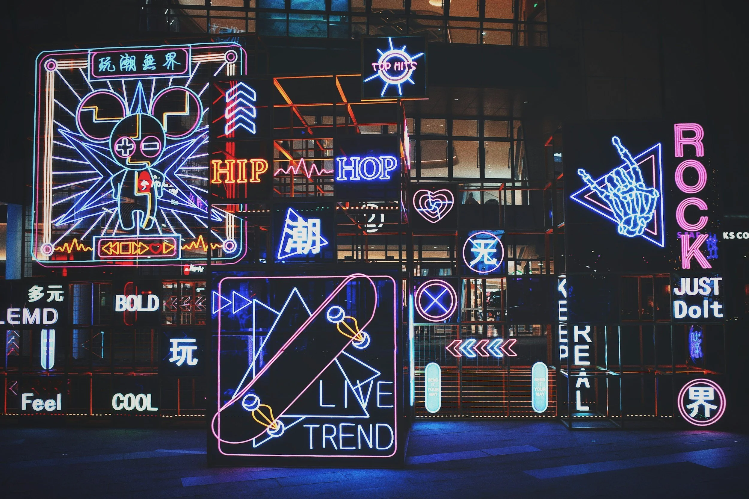 Colorful neon signs with various symbols, graphics, and words in English and Chinese. Features include a large sign with a character and Japanese characters, words like "HIP," "HOP," "ROCK," "LIVE TREND," and "JUST Do It," along with symbols such as a skateboard, a heart, a skull, and directional arrows, all set against a dark background.