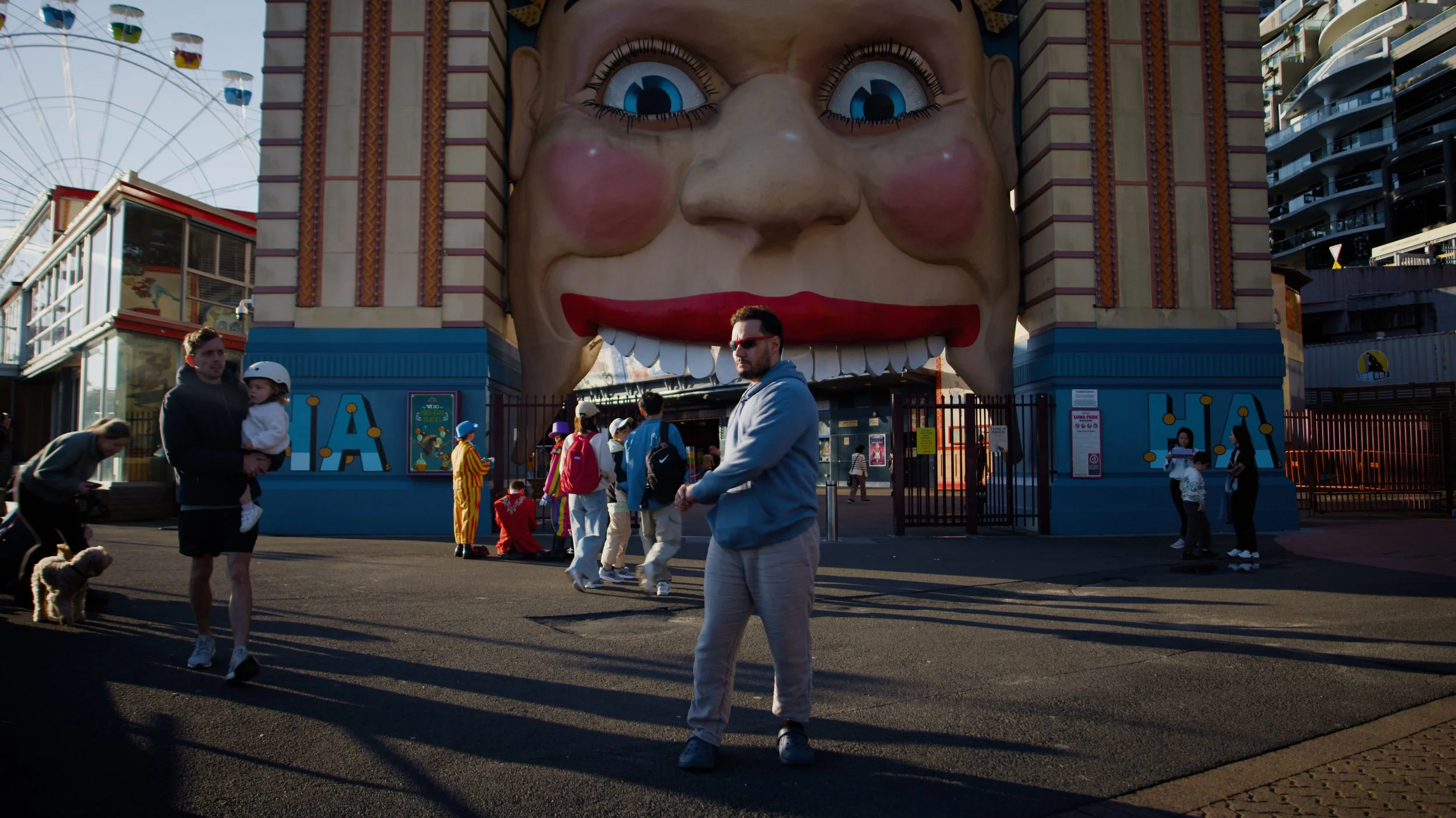 People walking outside of a carnival with a giant clown face entrance, including a man with sunglasses in the foreground and a man holding a child.