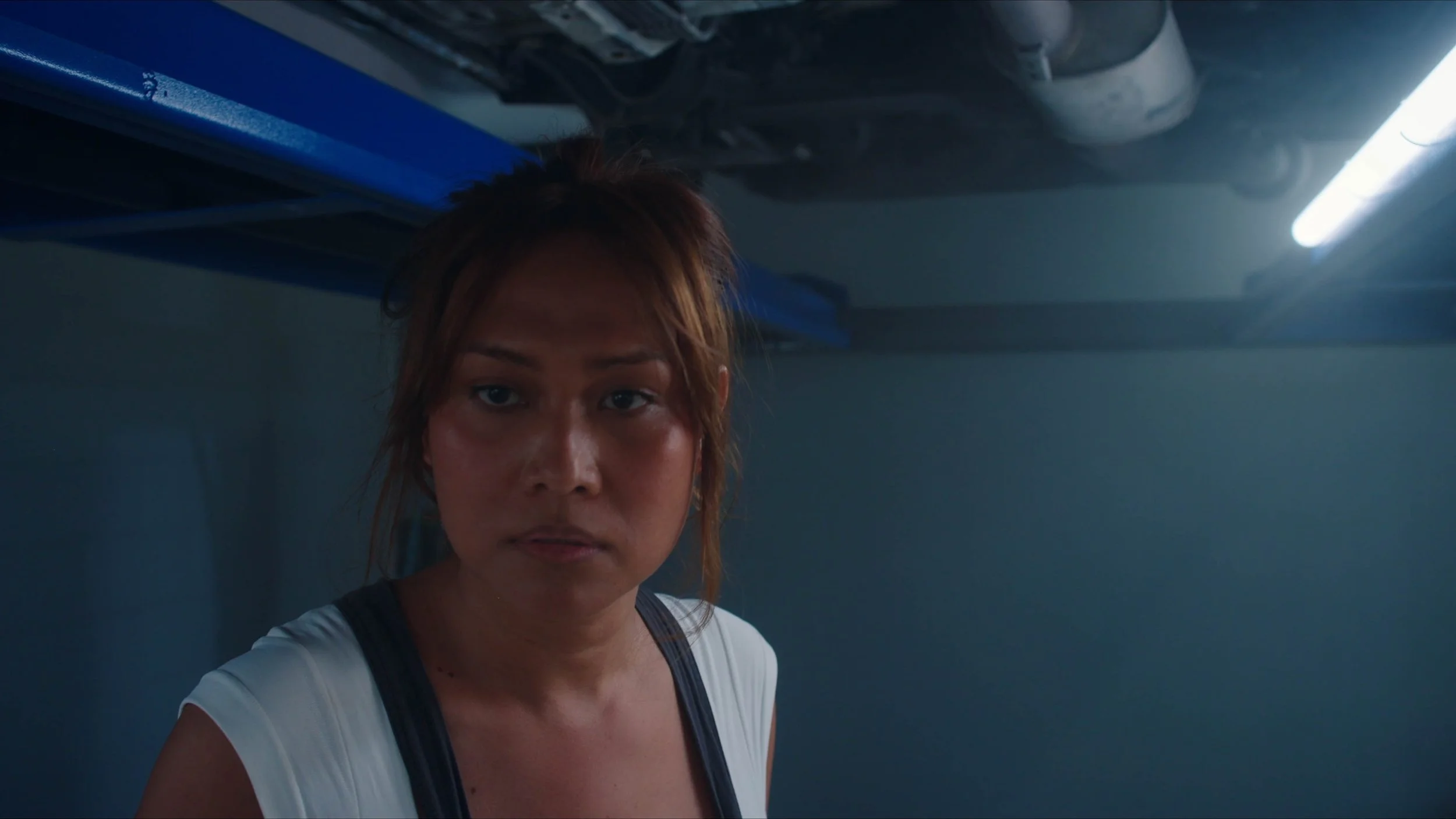 A woman with brown hair looking at the camera in a dimly lit space with a blue overhead object and a fluorescent light.