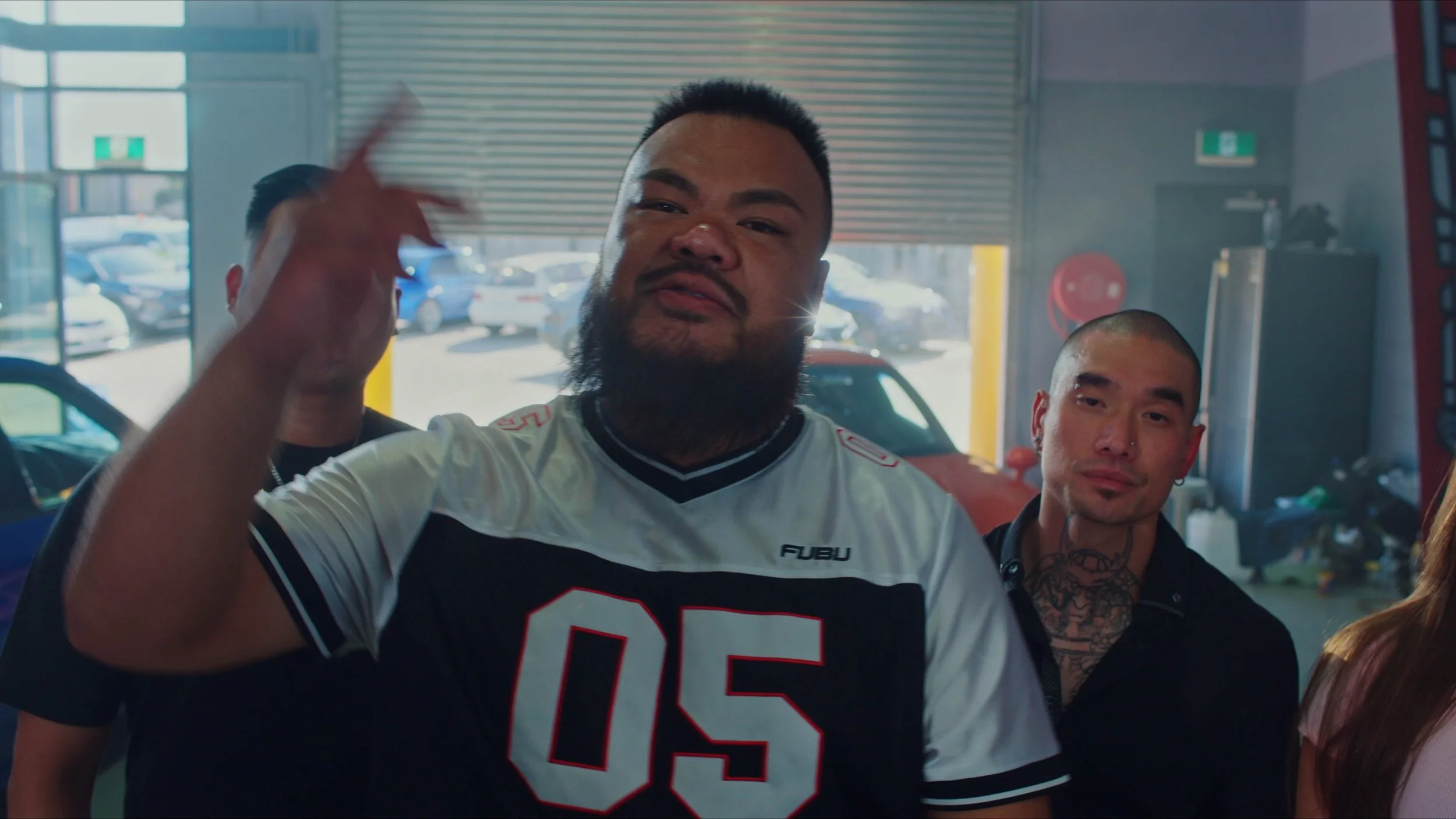 Three men in a car repair shop, with one in the foreground wearing a white and black sports jersey with the number 05, and two others behind him, one with tattoos and a shaved head, and the other partially visible.