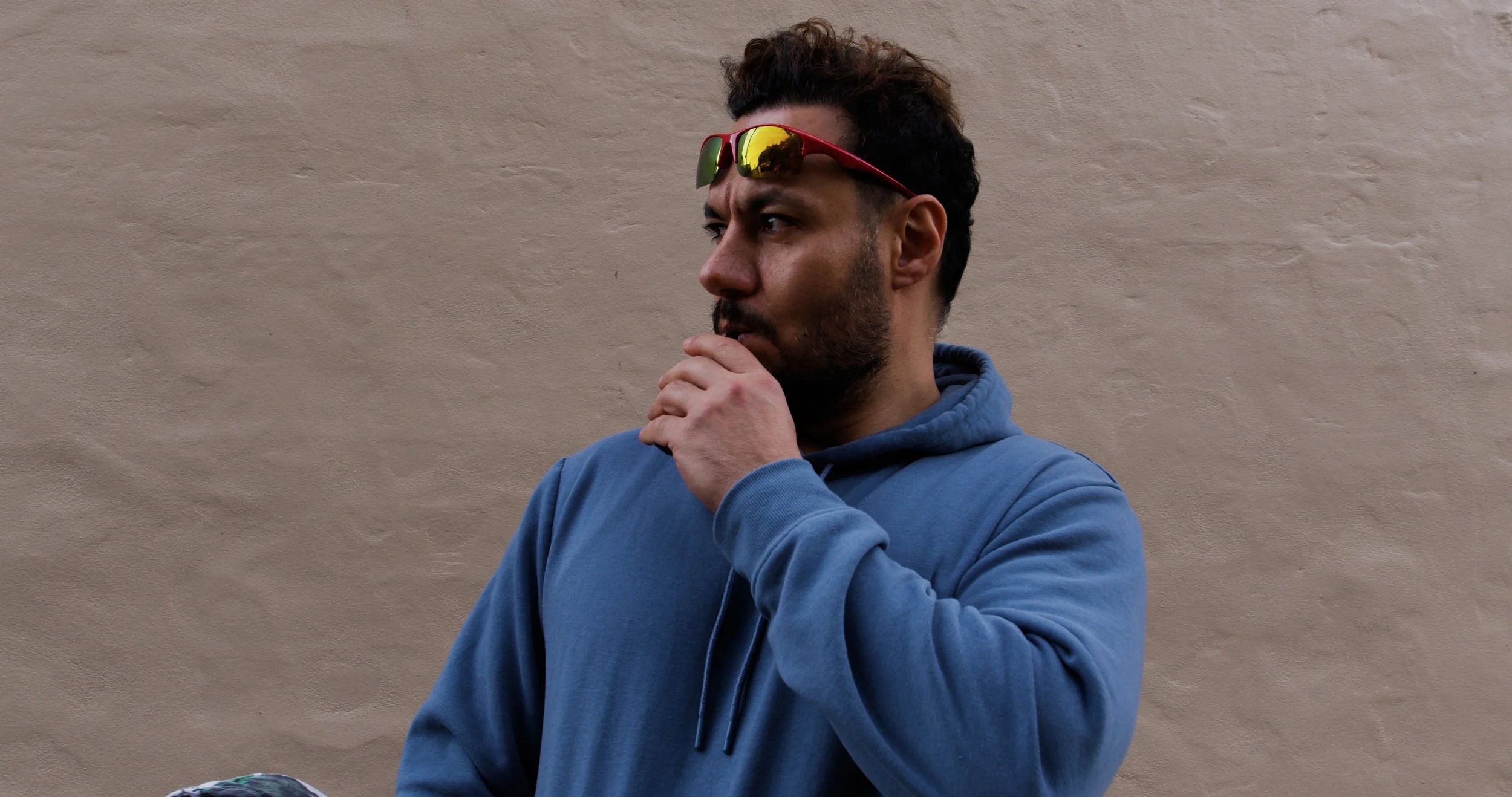A man with dark curly hair, a beard, wearing a blue hoodie and red sunglasses with reflective lenses, standing against a beige wall and touching his chin thoughtfully.