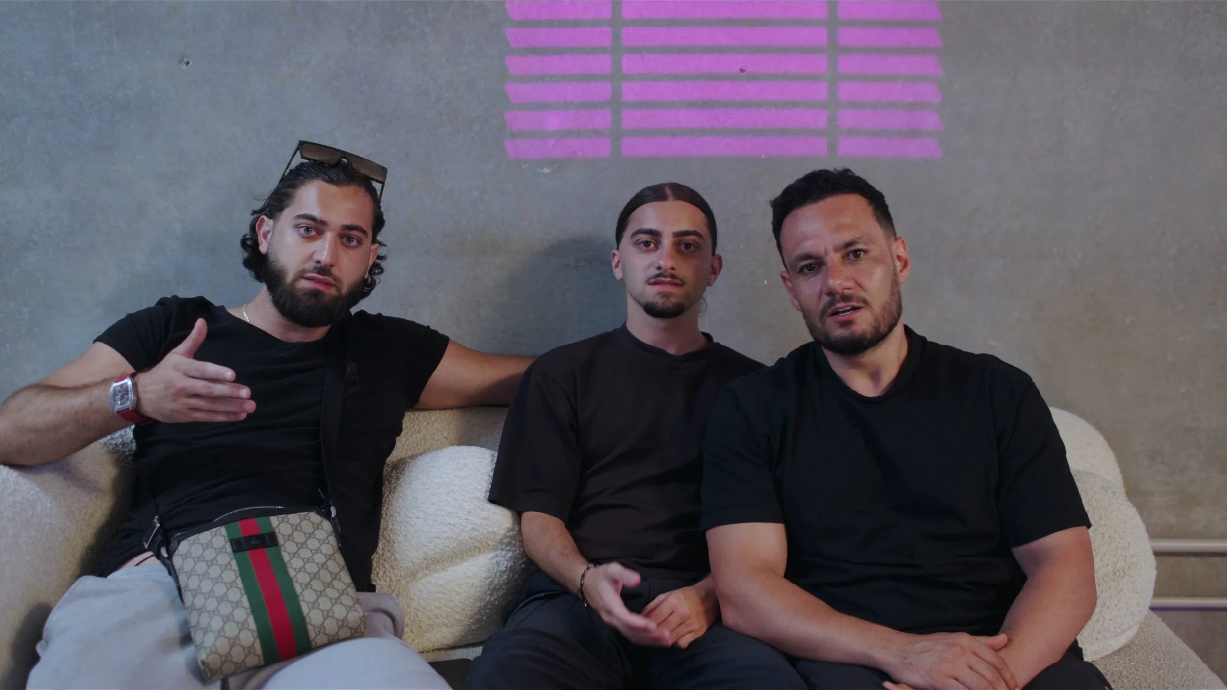 Three men sitting on a beige couch in front of a gray wall with pink stripes, dressed in black shirts, looking at the camera