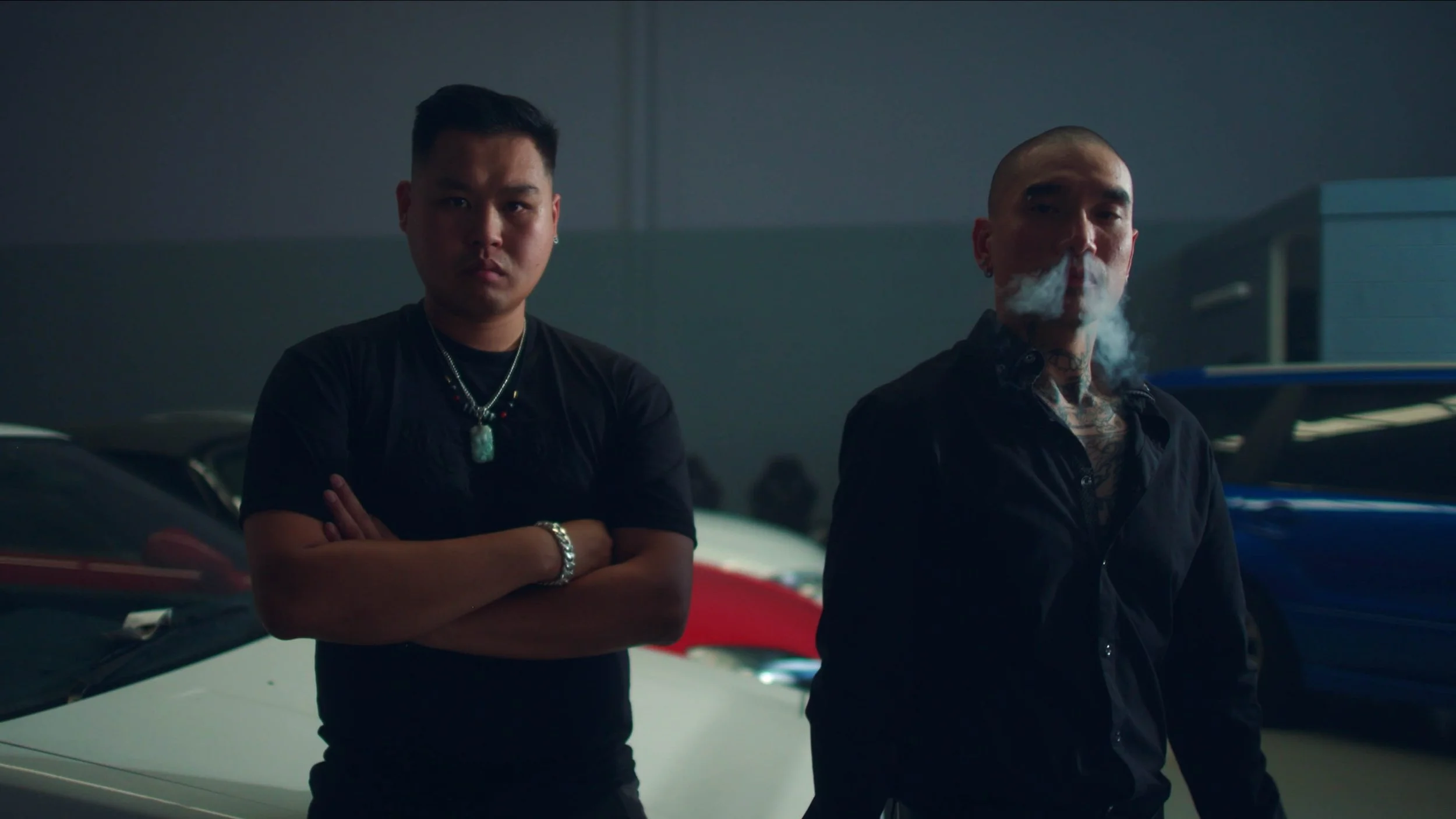 Two men standing in a parking garage, one with arms crossed and the other smoking, with cars in the background.