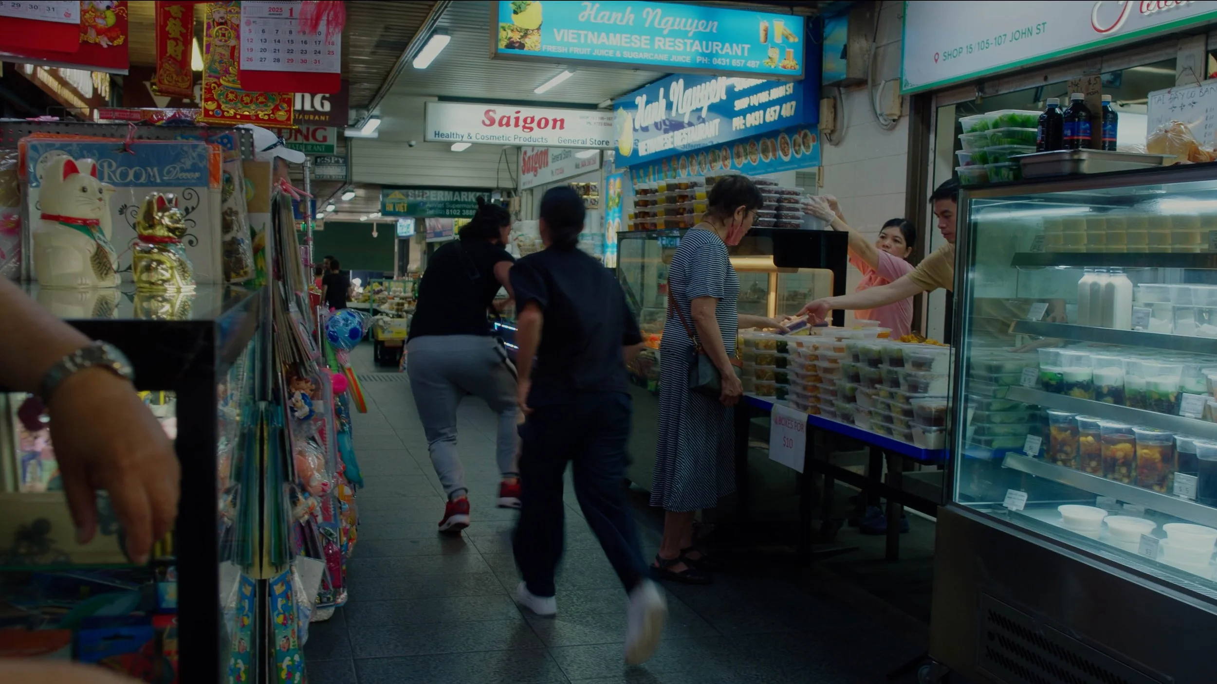 People shopping in an Asian market with food stalls and souvenirs, including ceramic cat figurines, signs in Vietnamese, and a variety of packaged food items.