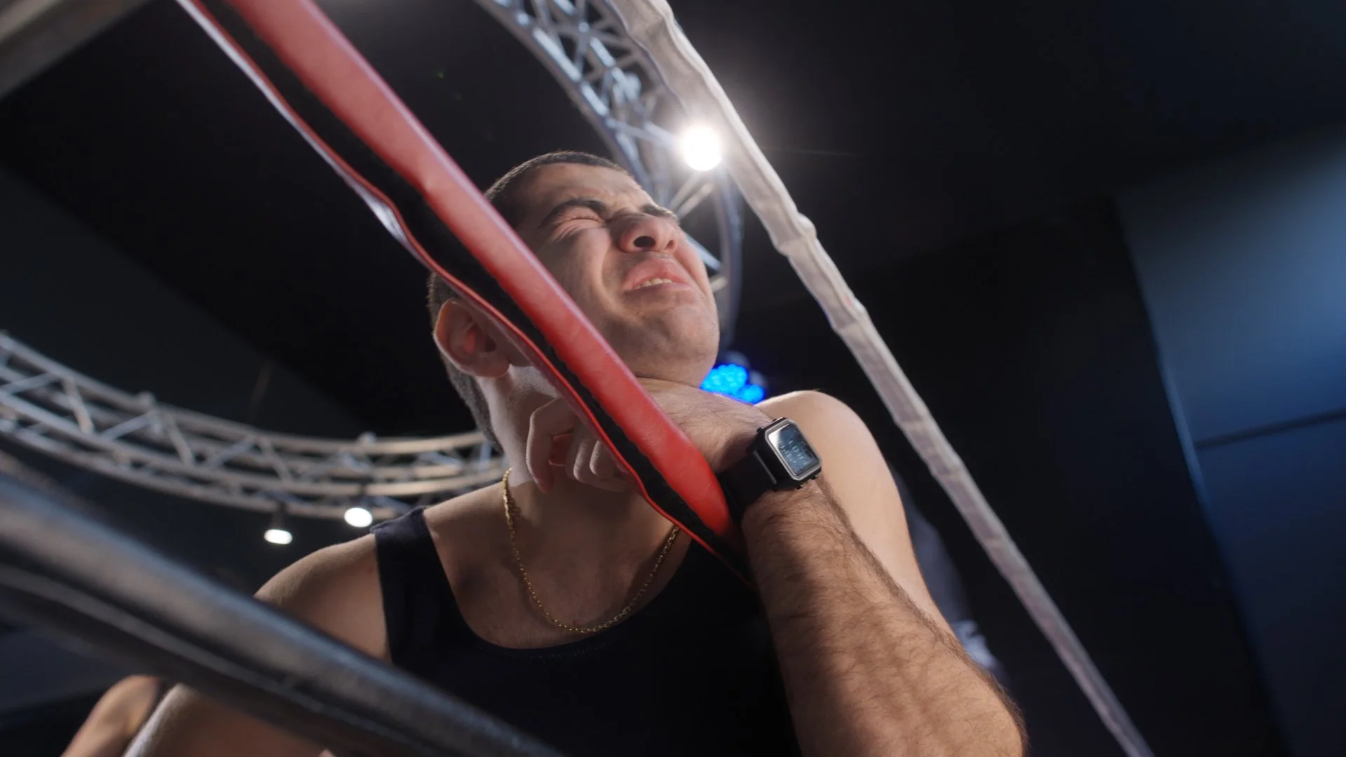 A boxer is grimacing in pain as a punching pad hits his neck during a boxing training session inside a gym with professional lighting and metal truss.