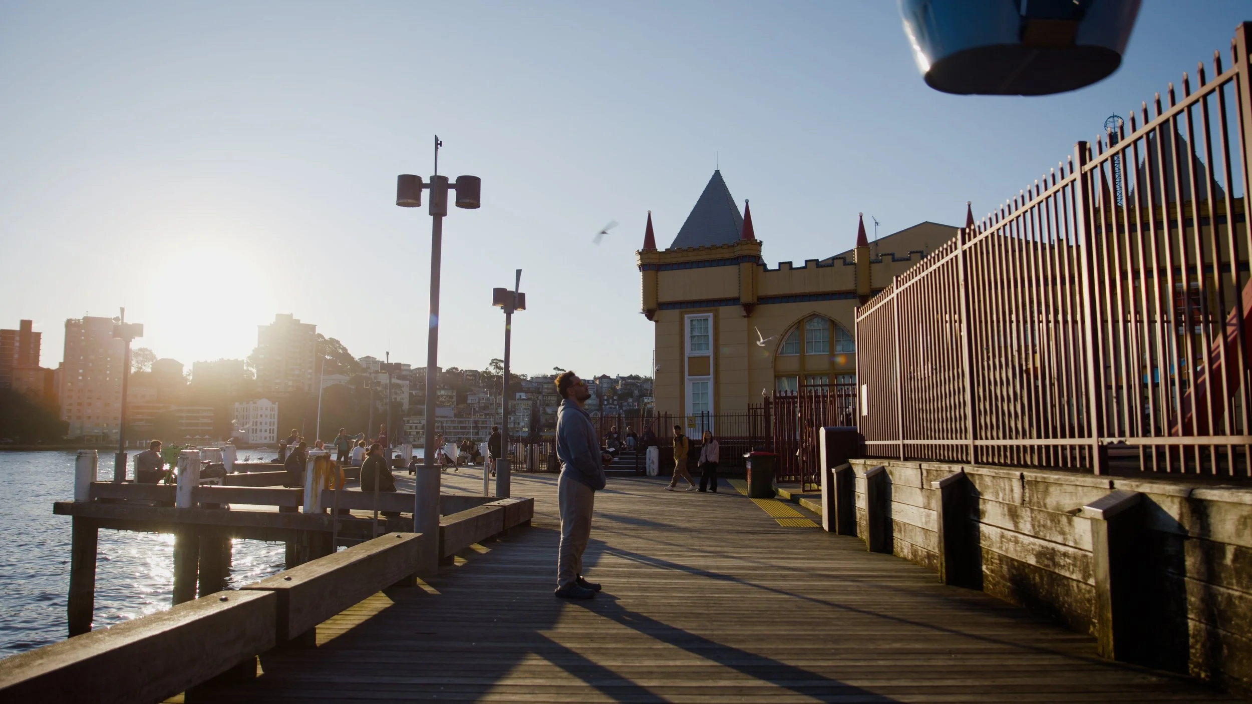 People on a wooden pier near a castle-like building with a fence, with the sun setting in the background and a few seagulls flying.