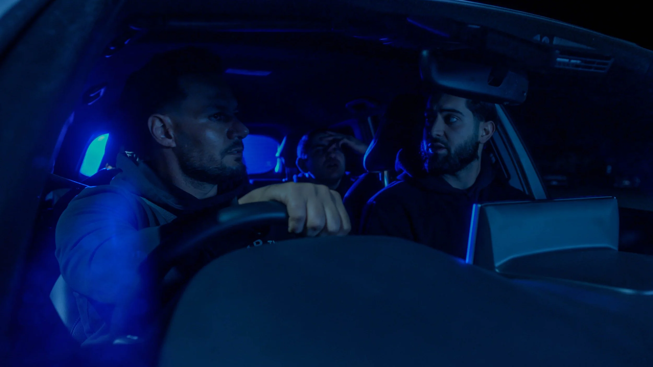 Three men sitting in a car at night, illuminated by blue interior lighting. The man in the front left is holding the steering wheel, the man in the front right is looking at him, and a third man is visible in the back seat making an expressive face.