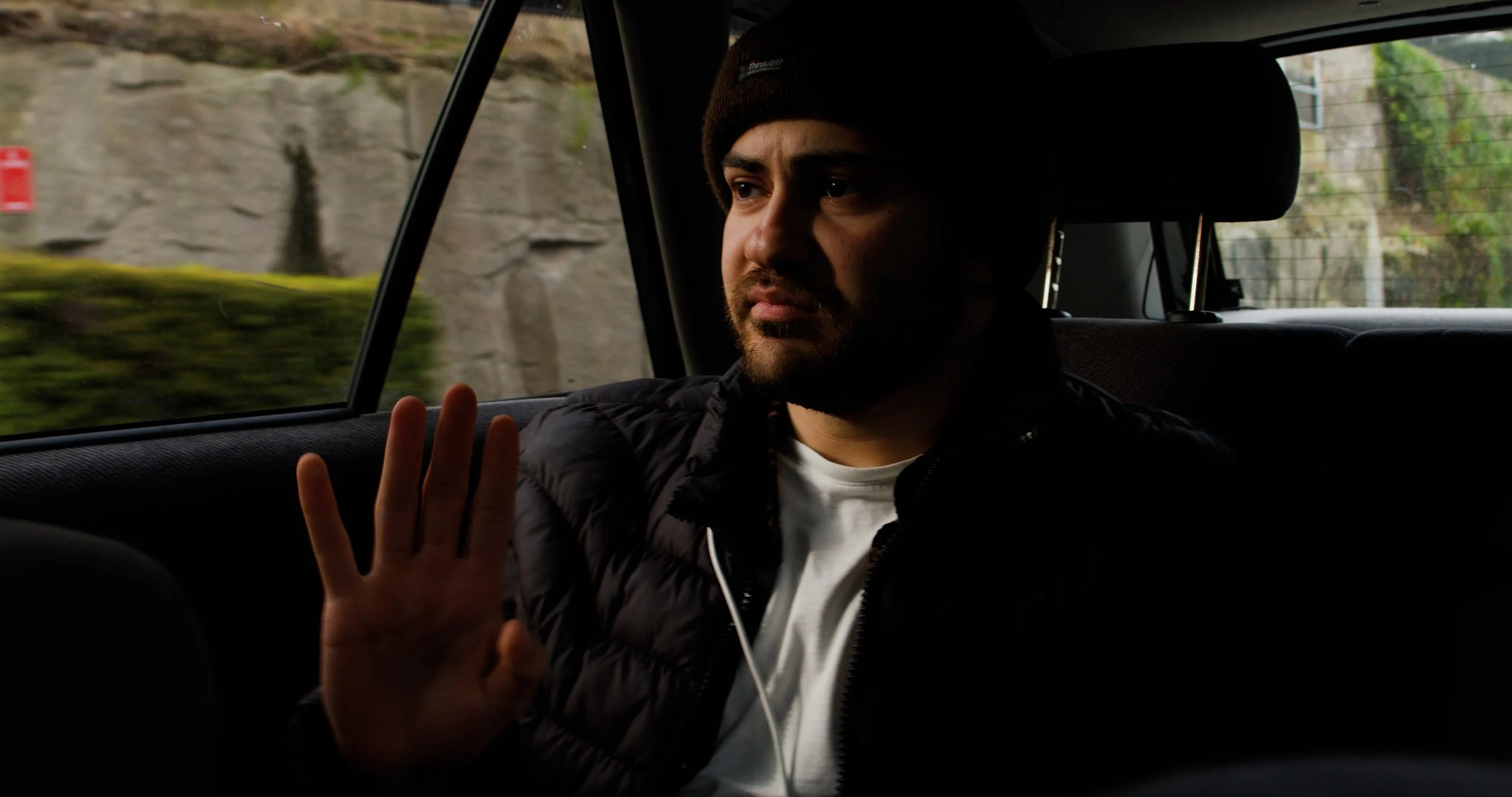 A man with a beard and brown beanie sitting in a car, facing sideways, with his hand raised in a gesture of stop or attention, with a serious expression.