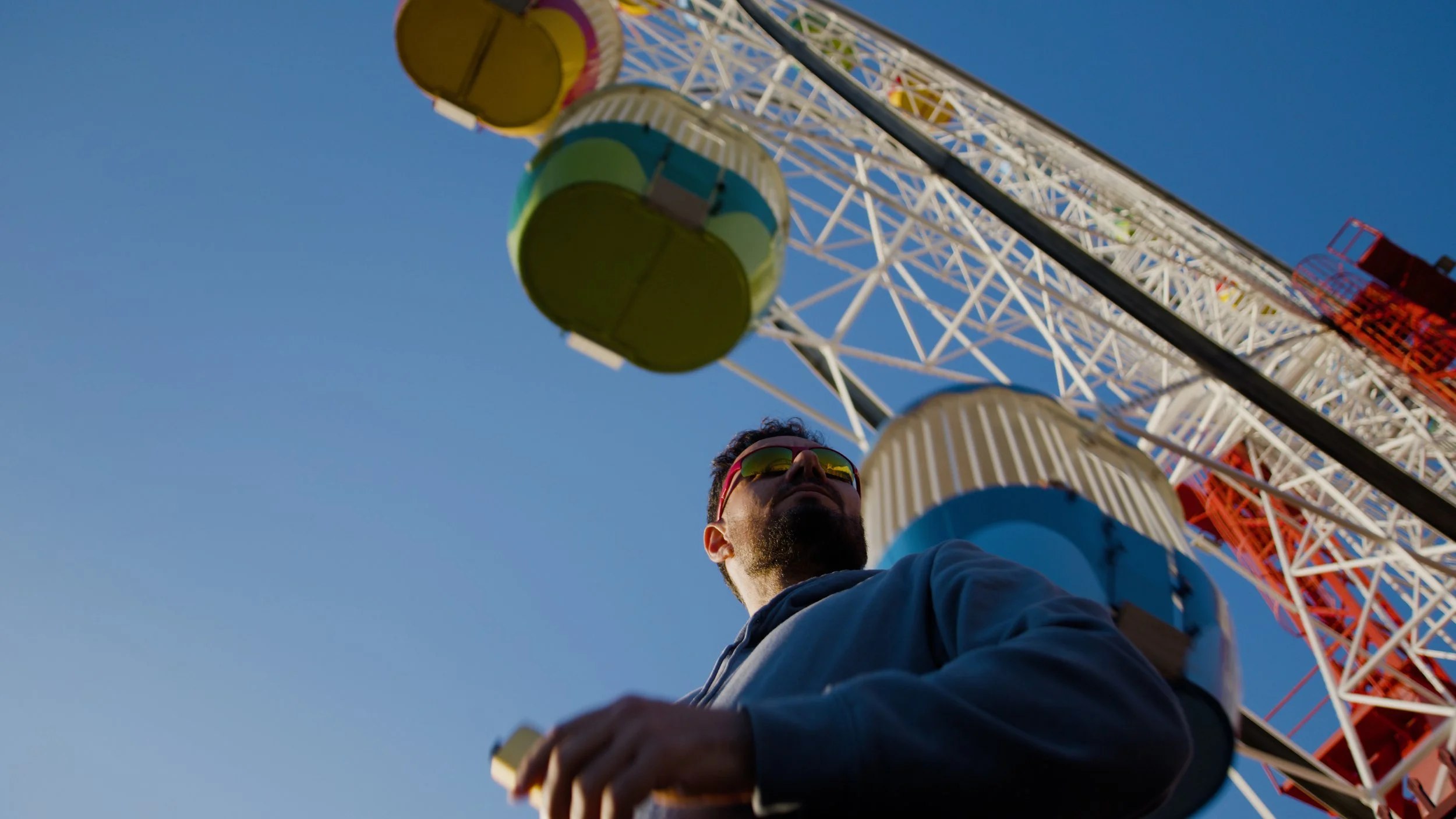 A person wearing sunglasses and a hoodie standing near a tall amusement park ferris wheel with colorful cabins against a blue sky.