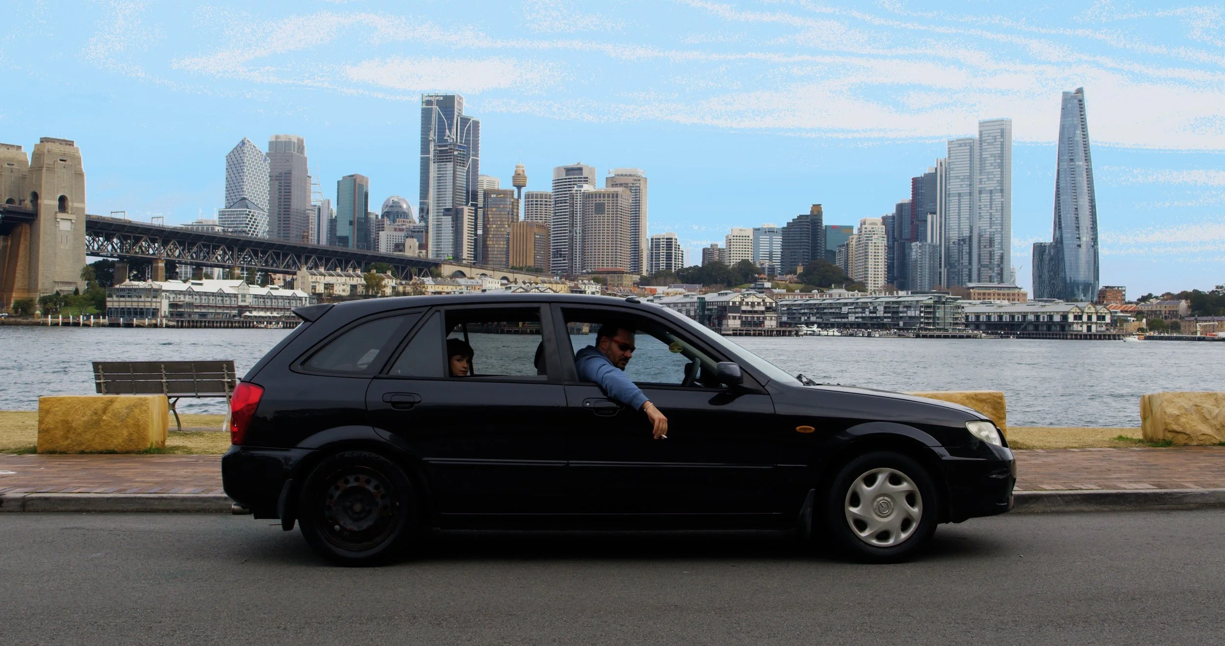 A black car parked along a waterfront with a city skyline in the background, including various tall buildings and skyscrapers.