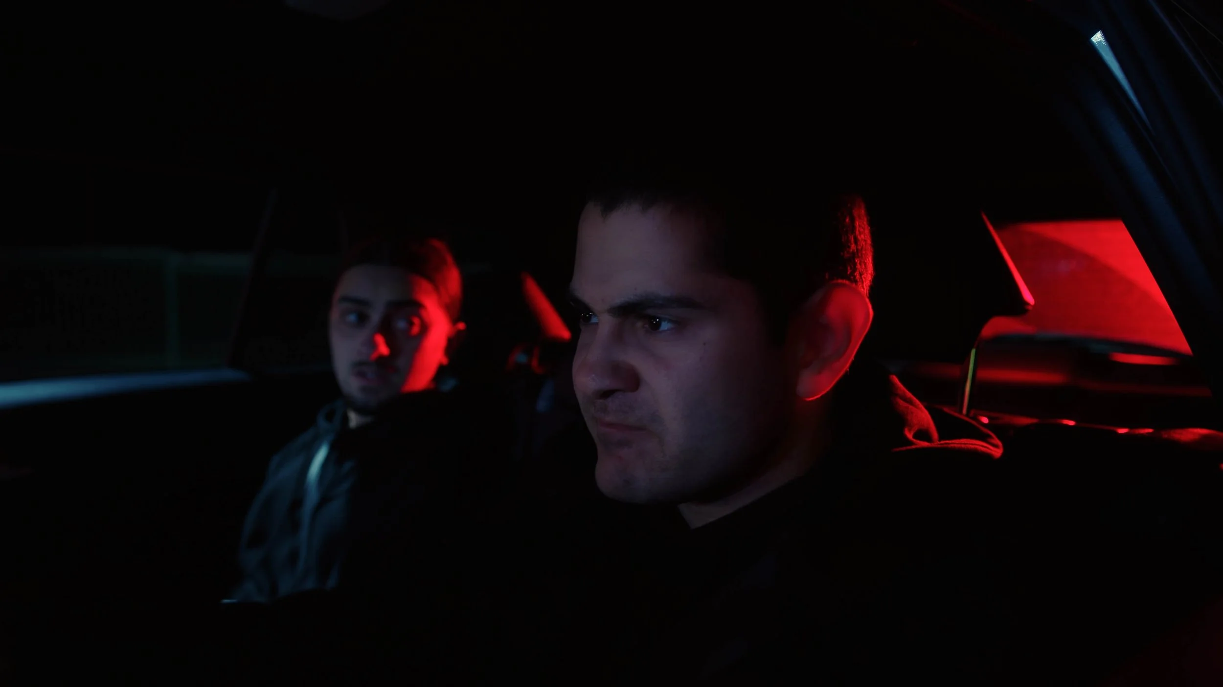 Two young men sitting in the backseat of a car at night, illuminated by red and dark lighting.