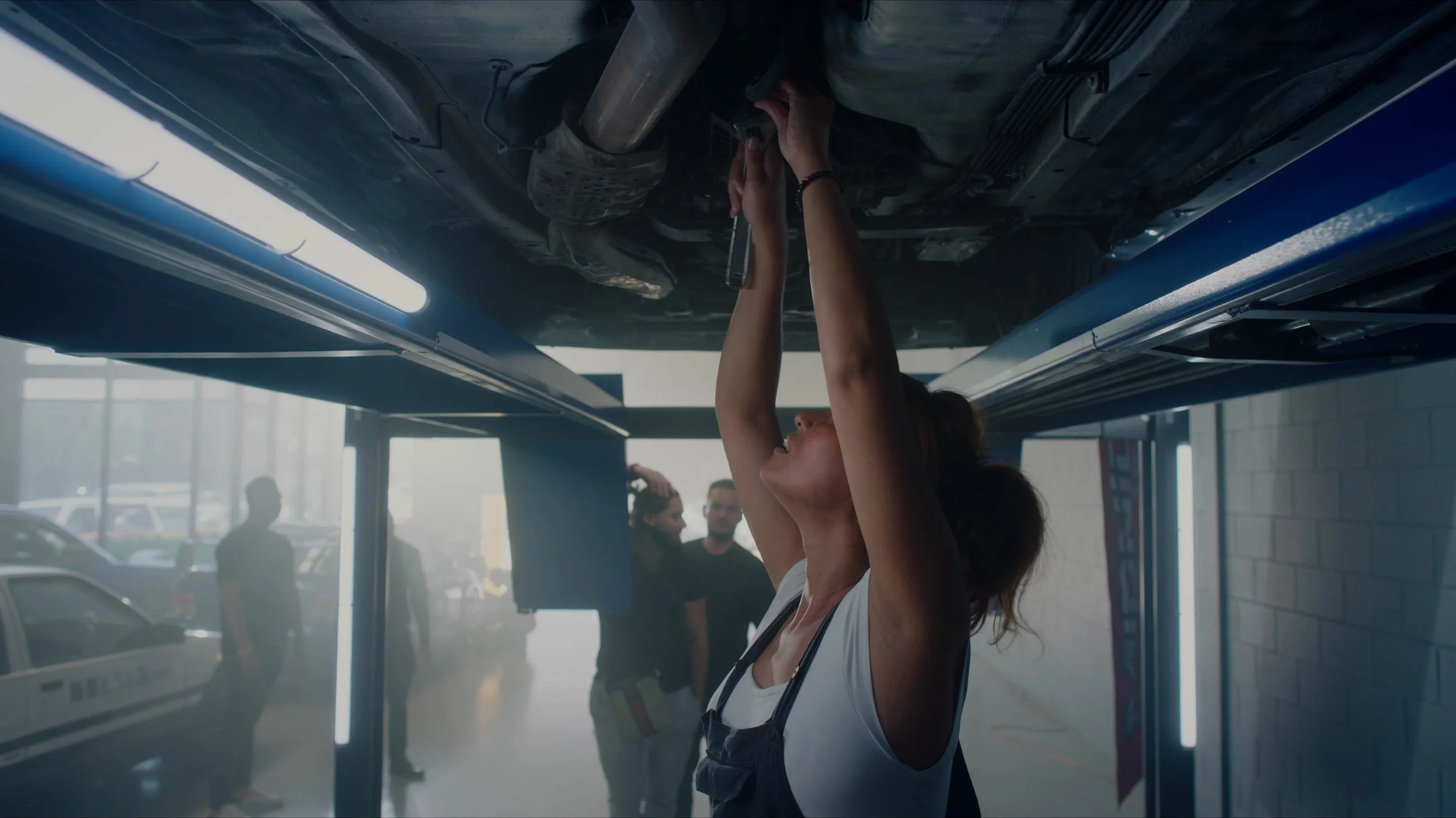 A woman working underneath a lifted car, using a wrench, in a garage, with other people in the background.