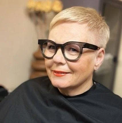 Kvinne med kort blond hår, iført svarte briller og rød leppestift, smilende.