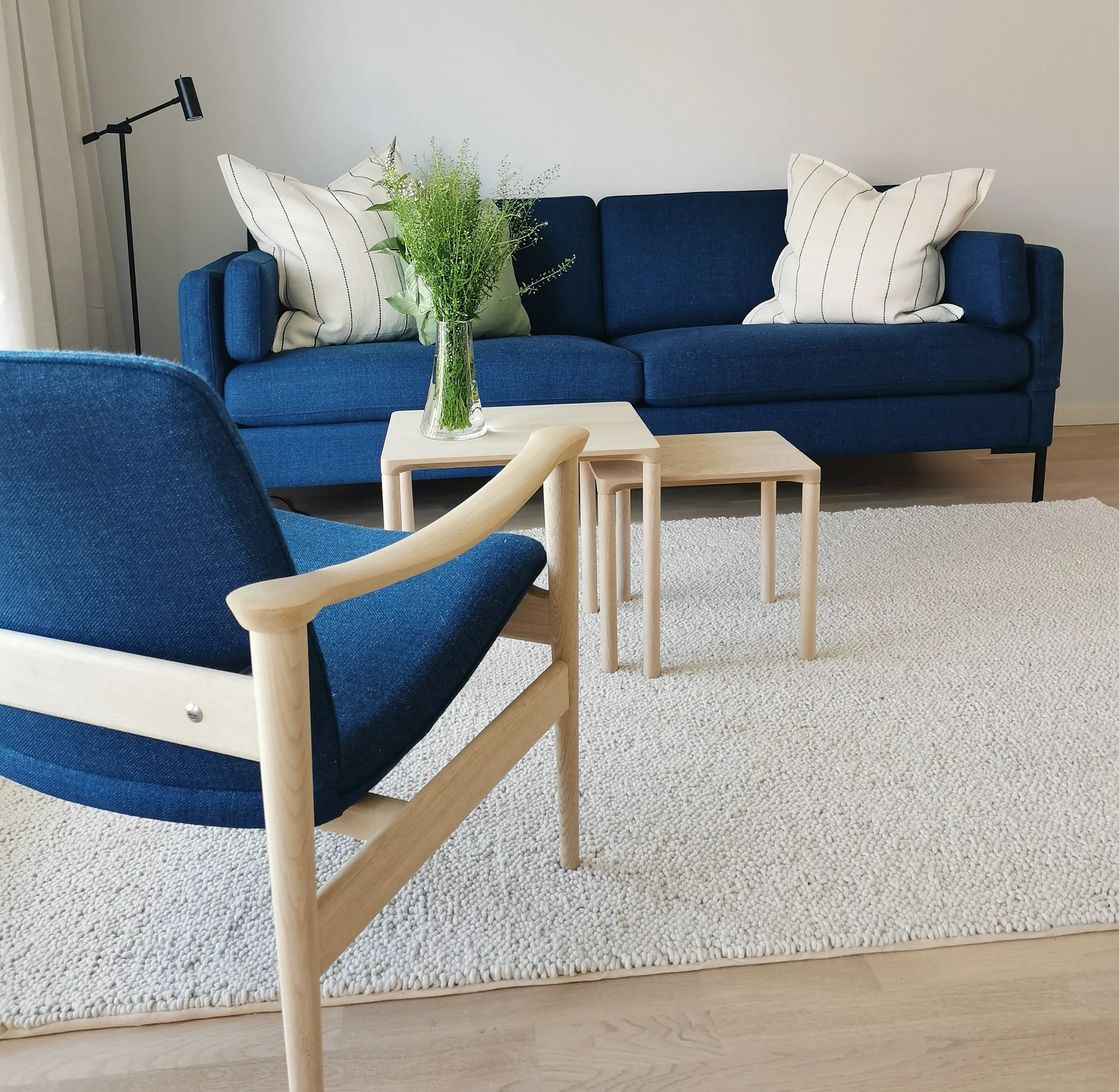Stue med blå sofa, to trekantede puter, et bord med en vase med grønne planter, en blå stol med tre små sidebord, og en hvit teppe på lys treparkett.