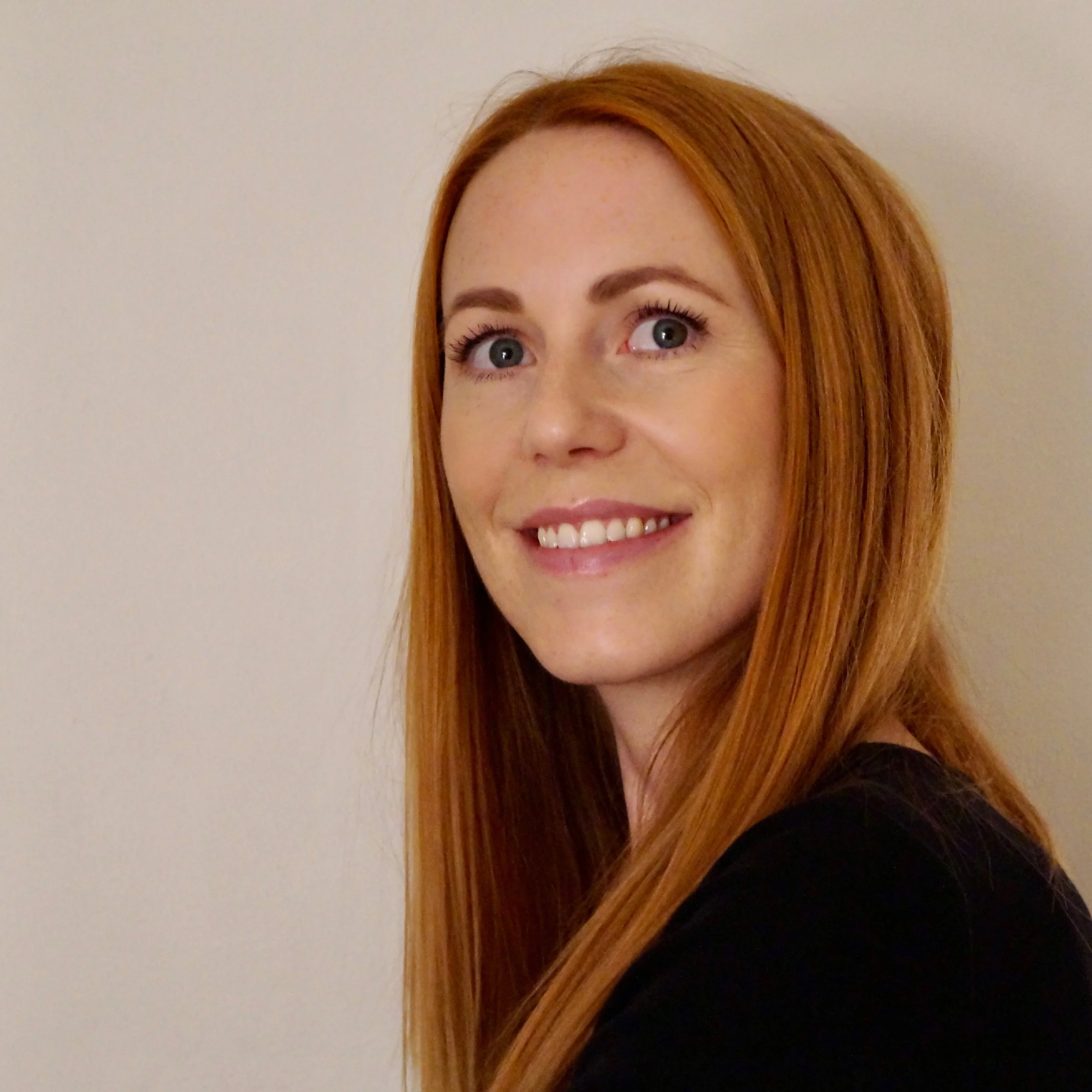 Portrett av en smilende kvinne med langt, rødt hår, iført svart, foran en lys vegg.