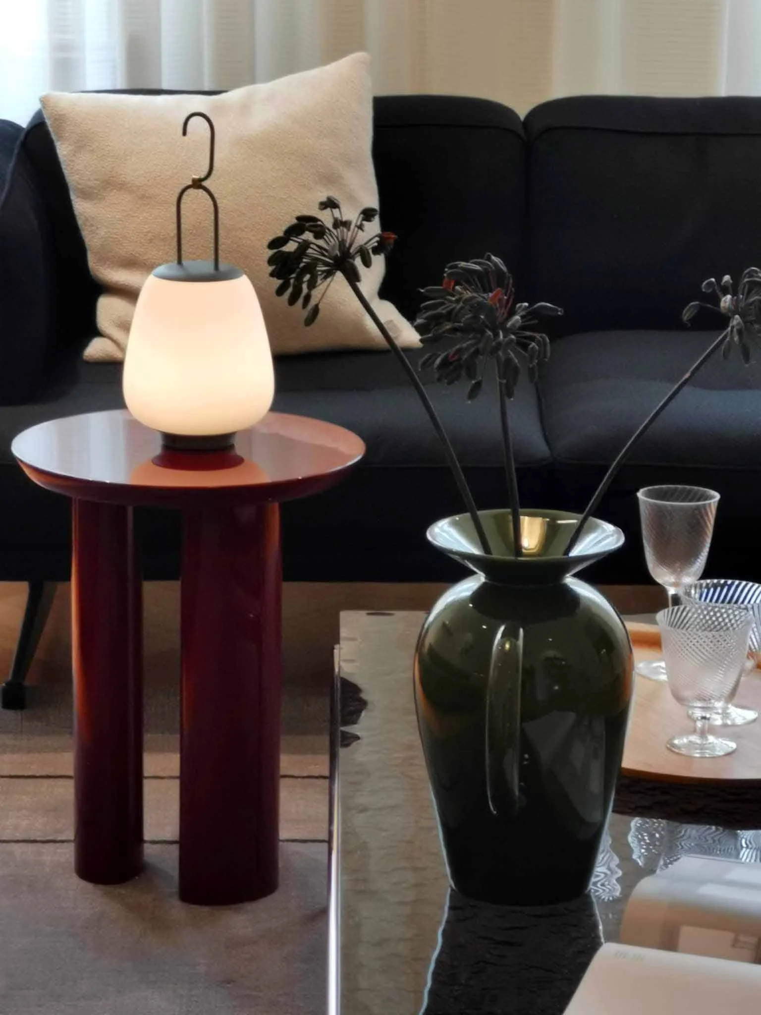 Et svart sofa med en beige pute foran et kuddfotsted med en lampe og en grønn vase med blomster.
