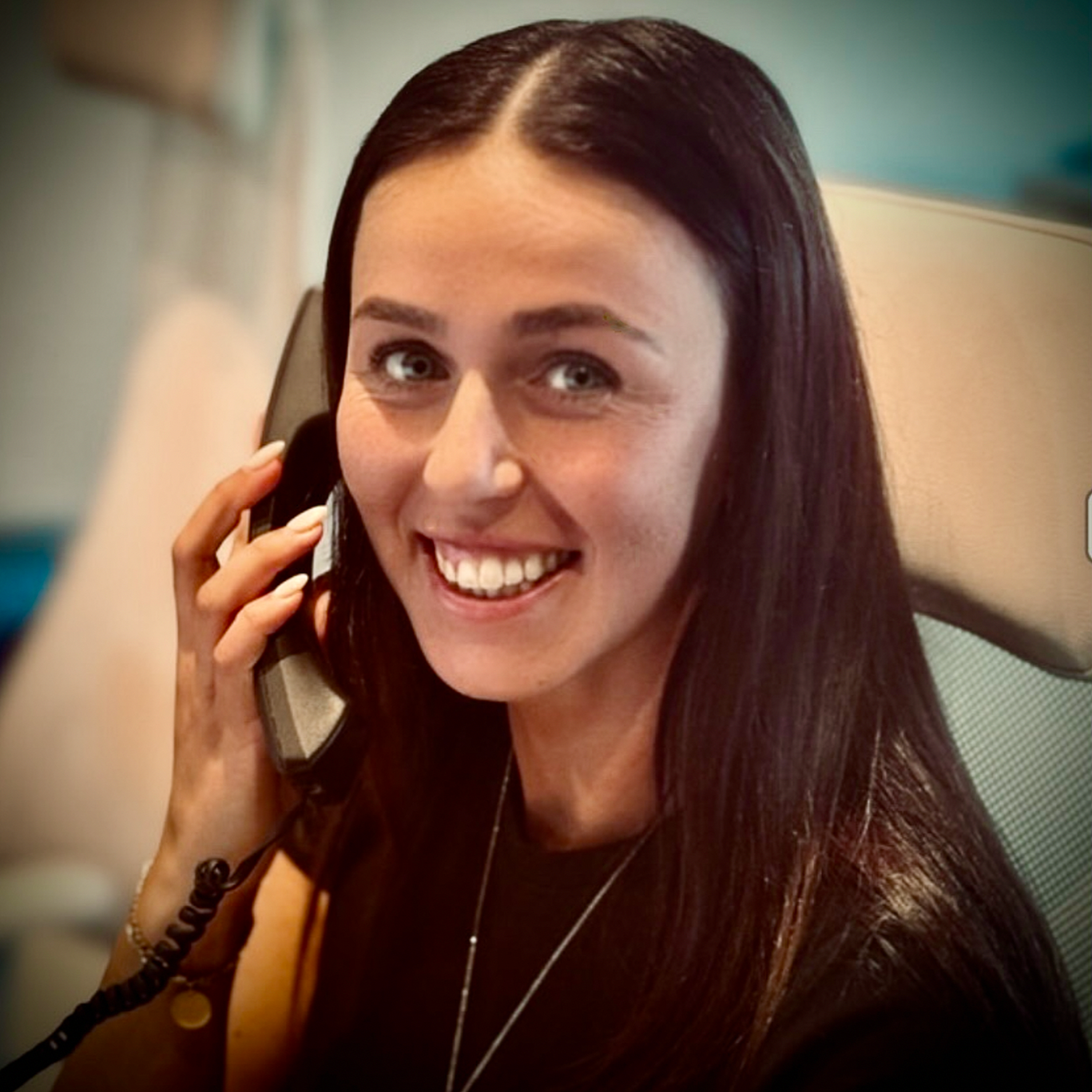 A woman with long dark hair smiling and talking on a landline phone in an office environment.