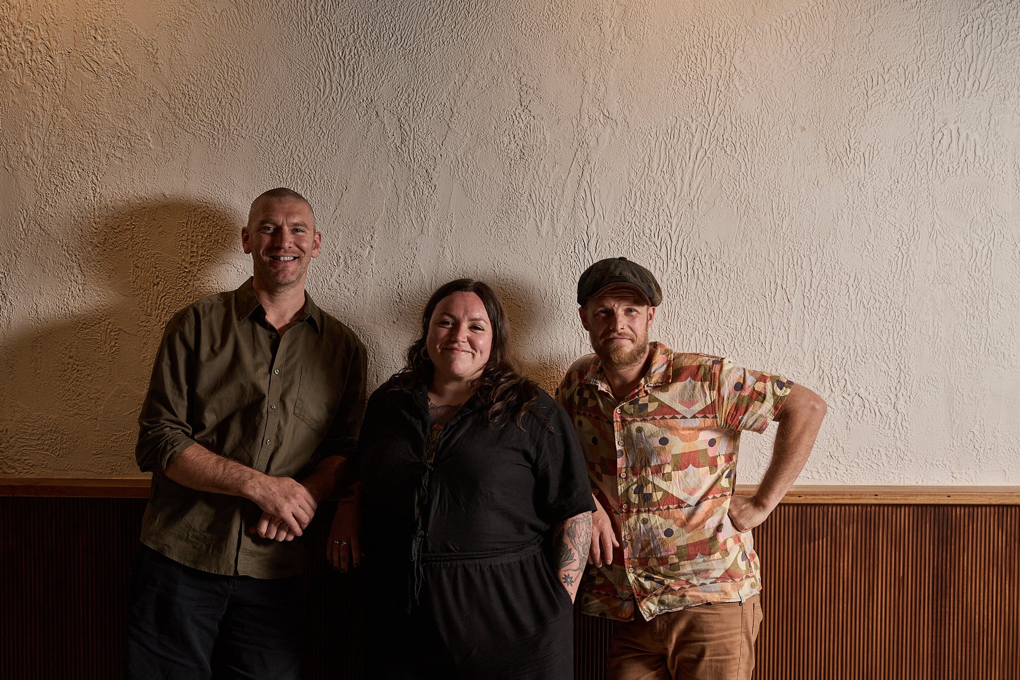Three people standing in front of a beige textured wall with a wooden panel at the bottom. The person on the left is a man with a bald head, wearing a brown button-up shirt. The person in the middle is a woman with long dark hair, wearing a black jacket. The person on the right is a man with a beard, wearing a colorful patterned shirt and a cap. They are smiling and posing casually.