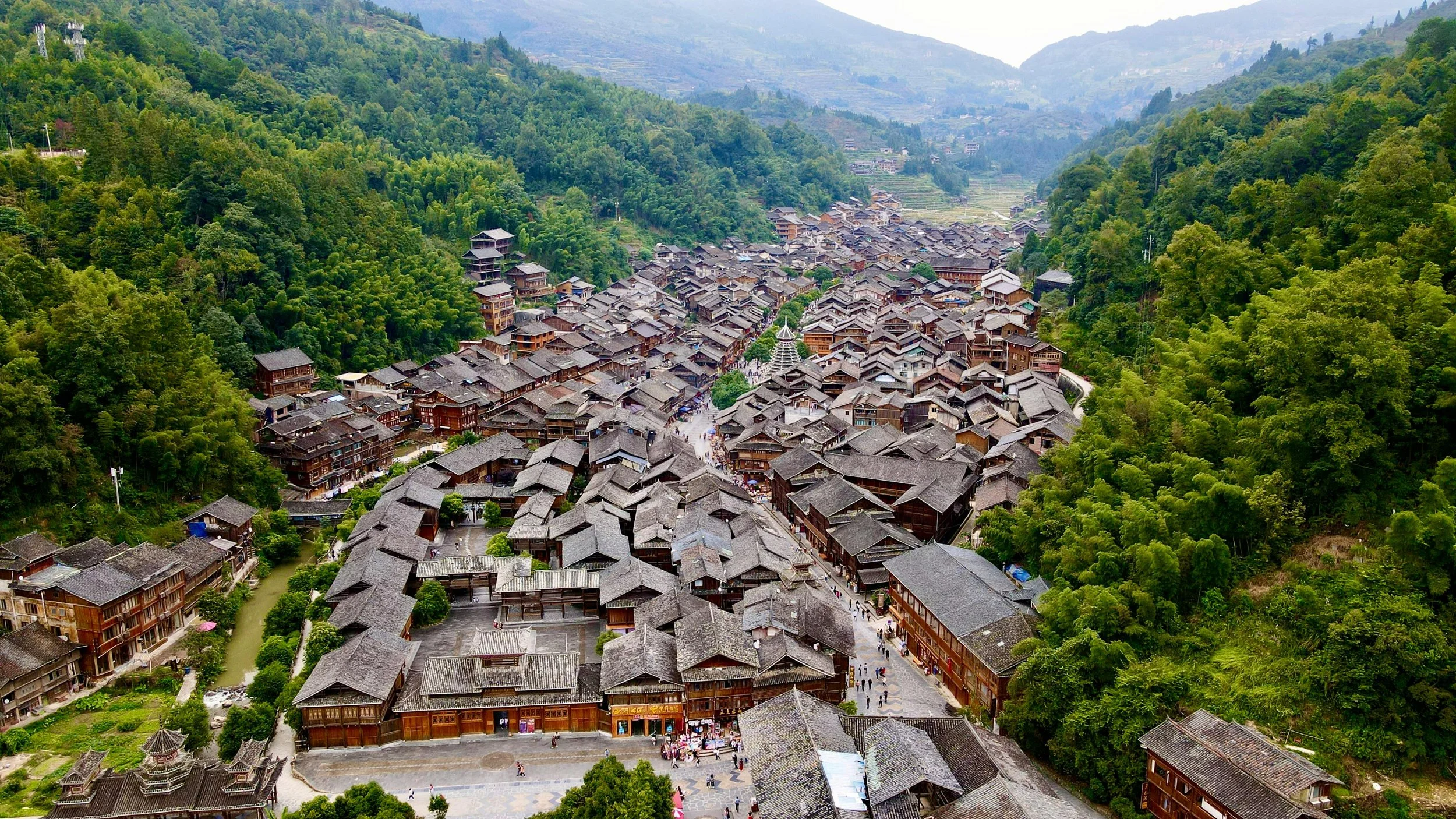A historic Miao ethnic village in Guizhou, China surrounded by green mountains with traditional wooden houses, narrow streets, and lush vegetation.