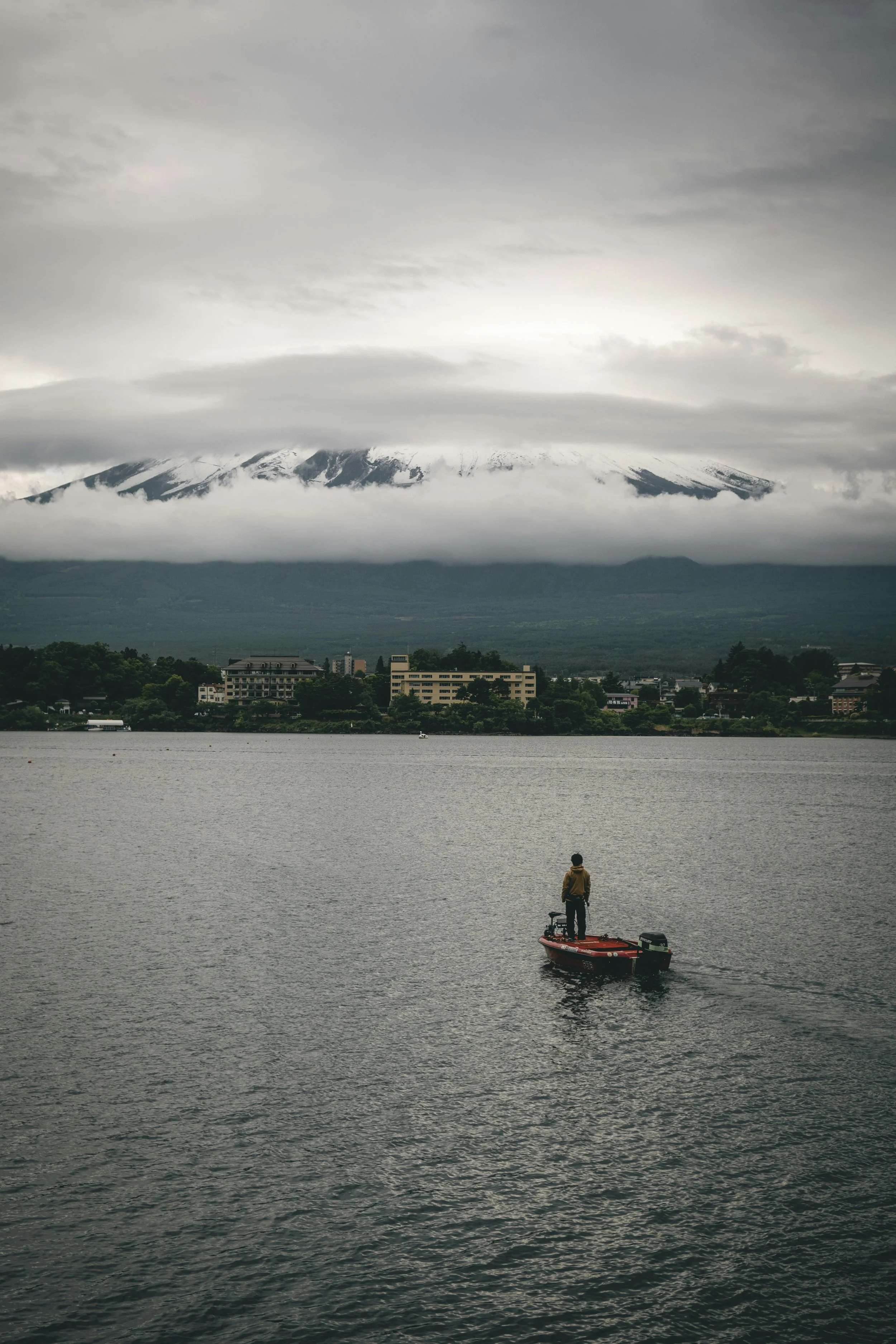 A person standing on a small boat on a lake in Japan, with a city and snow-capped mountain in the background, under a cloudy sky.