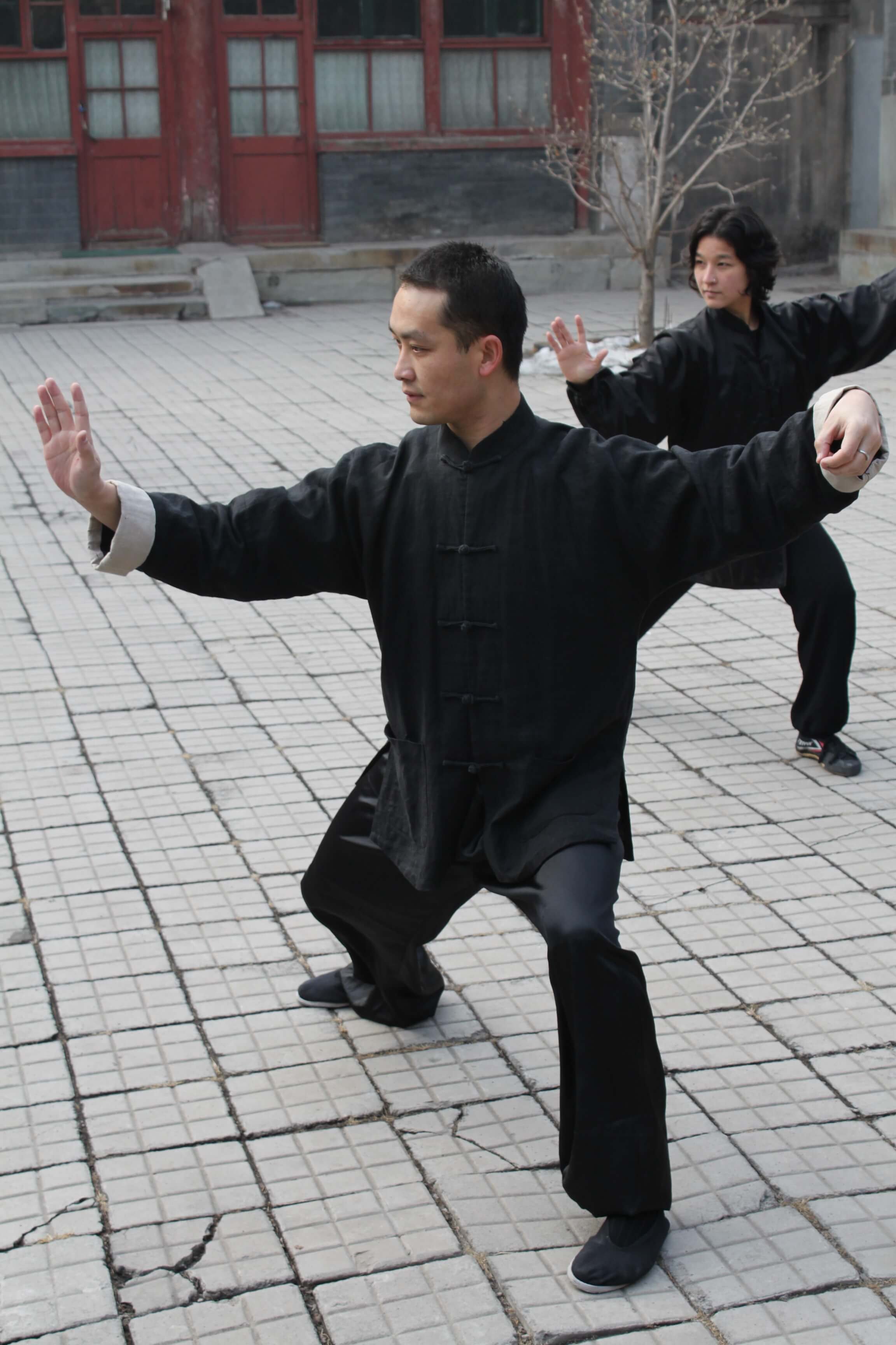 Two individuals practicing martial arts outdoors on a tiled courtyard, with traditional Chinese architecture in the background.