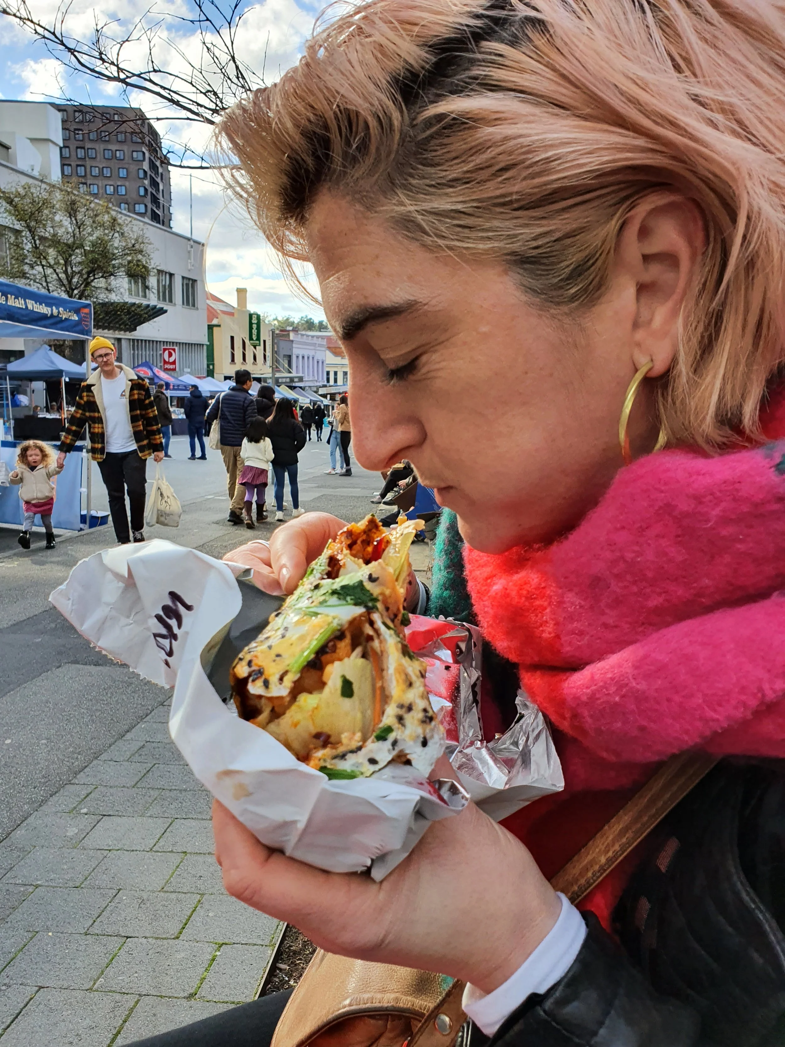 Woman with pink hair and gold hoop earrings eating a burrito on a busy outdoor street market.