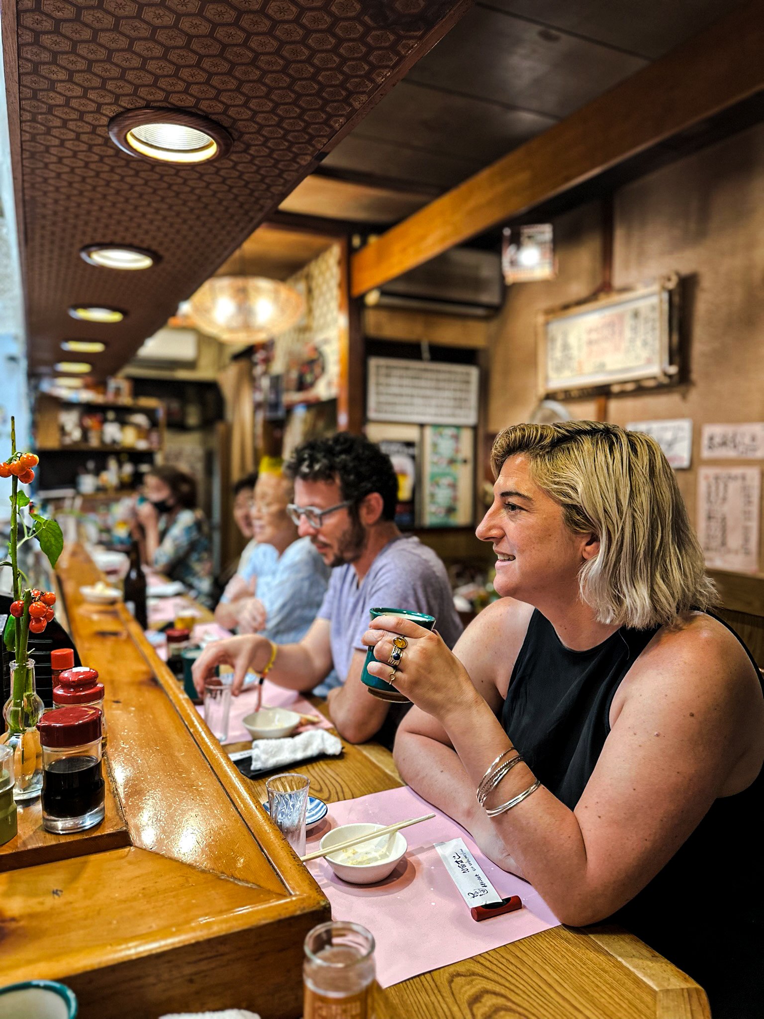 People sitting at a long wooden counter in a cozy sushi restaurant in Nagoya, enjoying drinks and conversation, with various condiments and small bowls on the counter.