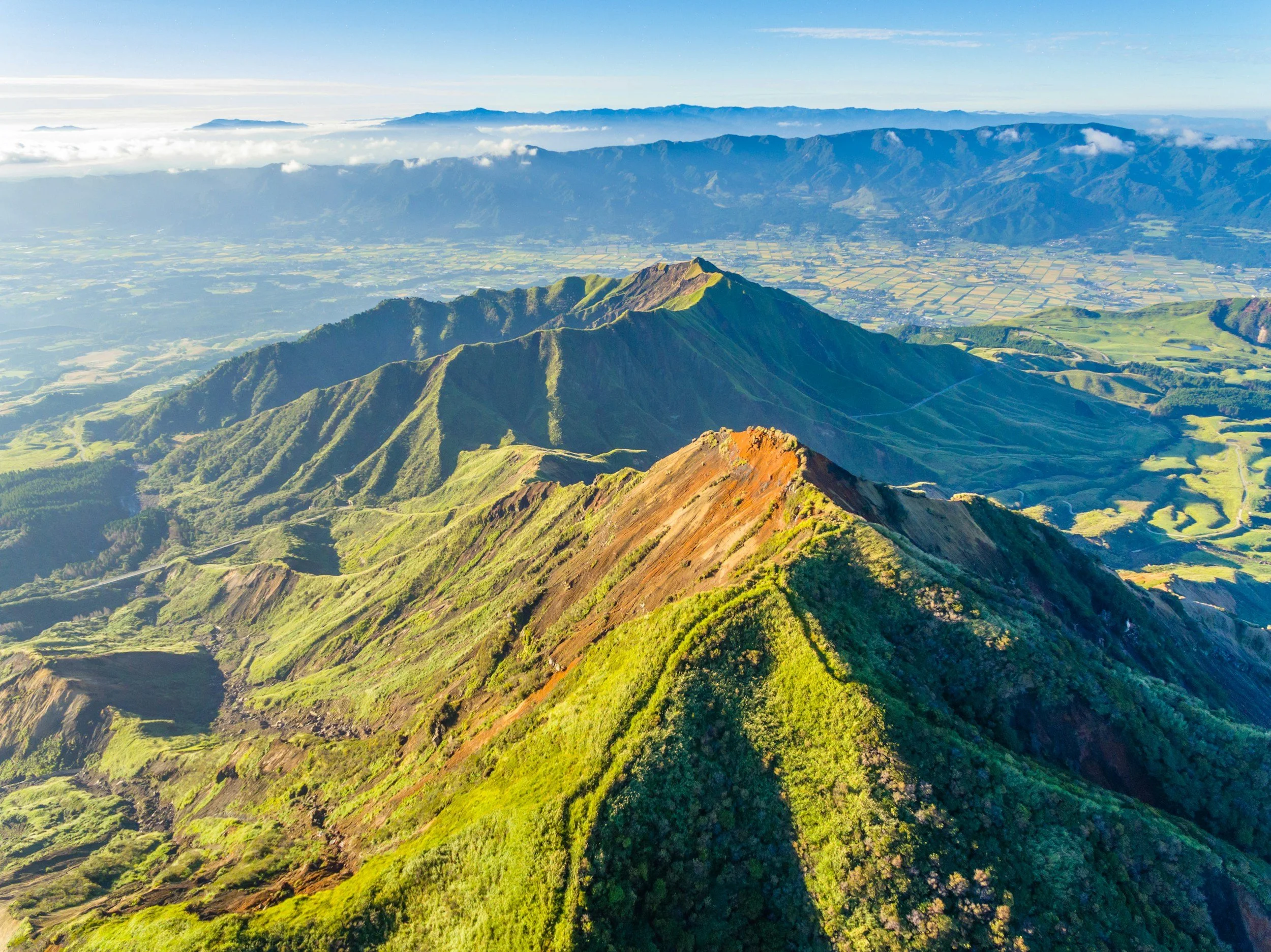 Aerial view of lush green Mount Aso in Kumamoto and valleys during daytime with a partly cloudy sky and distant mountain ranges in the background.