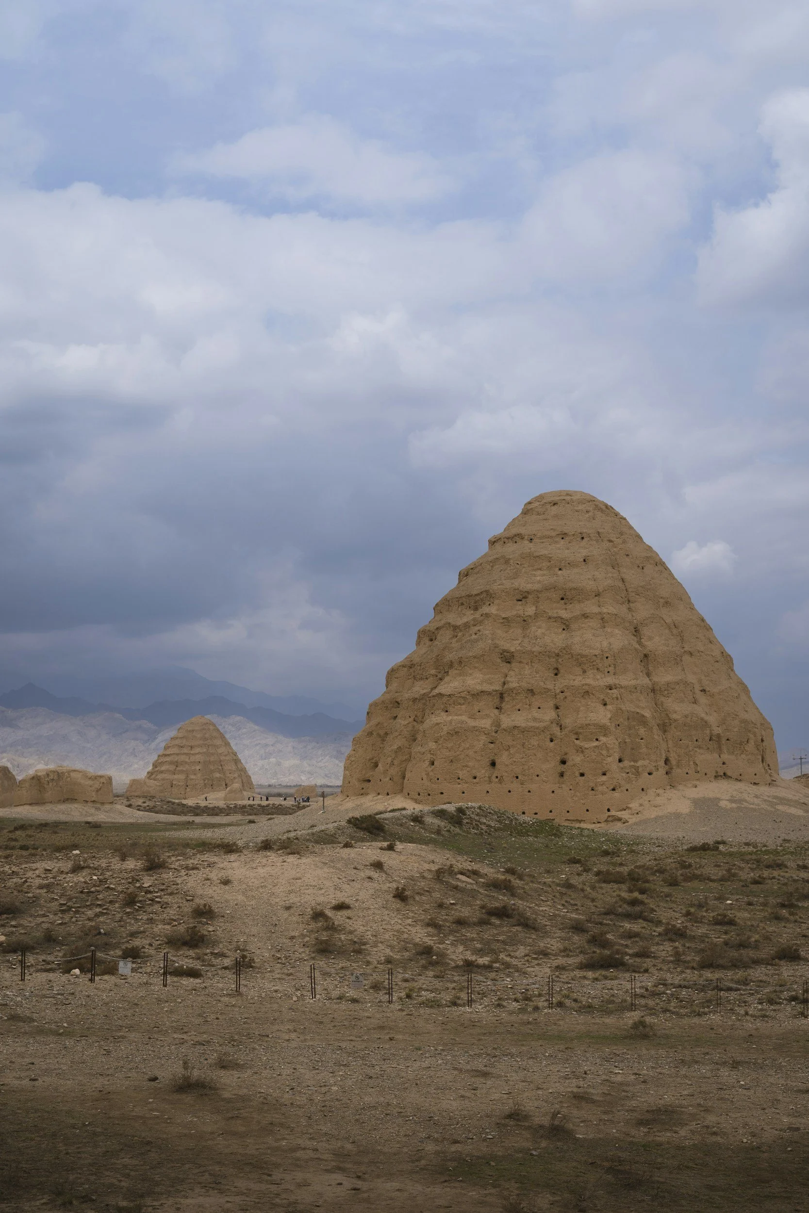 A desert landscape of the Xia Tombs in Ningxia with three large ancient mud-brick structures, possibly tombs or mausoleums, set against a cloudy sky with mountains in the distance.
