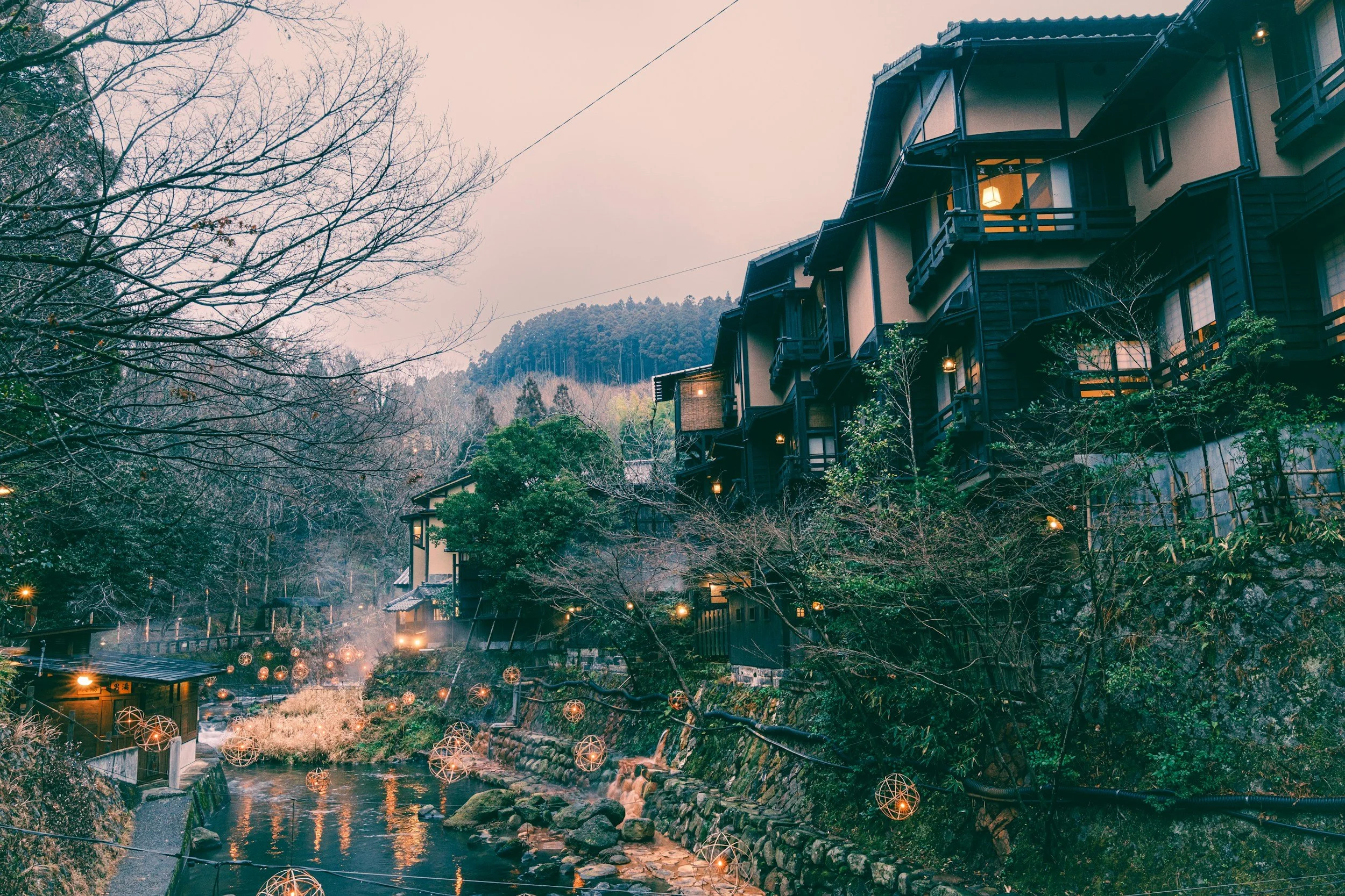 A scenic view of an onsen town in Japan with a river flowing through a village, with traditional houses on a hillside, surrounded by trees with some leaves fallen, and decorative string lights hanging over the river at dusk.