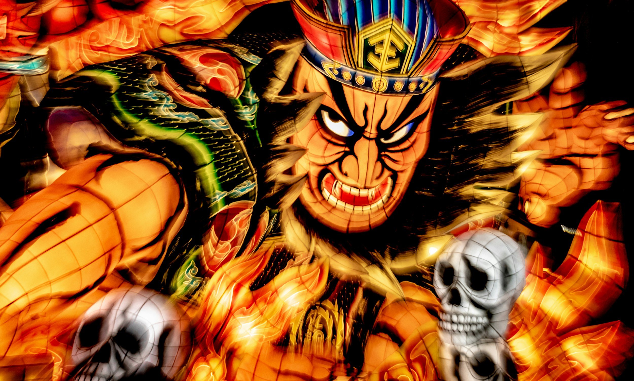 Colorful illuminated lantern of a fierce, angry warrior with blue eyes, horns, and a crown, accompanied by skulls, with a fiery background at the Nebuta matsuri festival in Aomori, Japan.