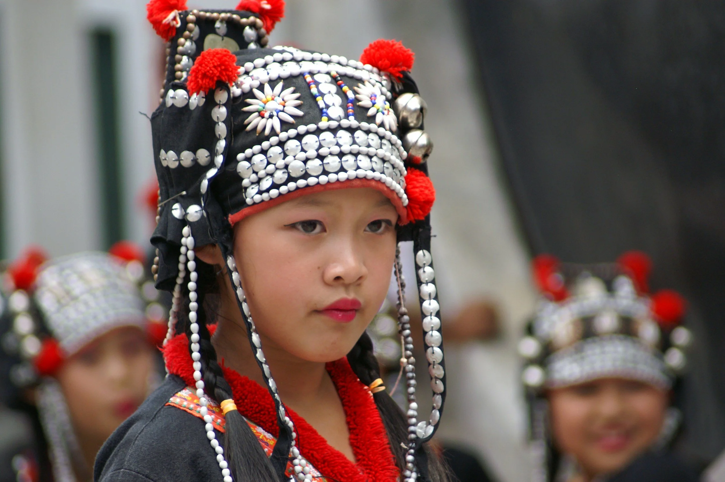 Young girl wearing traditional Tribal attire with a decorated headdress featuring beads, silver bells, red pompoms, and colorful embroidery, participating in a cultural event.