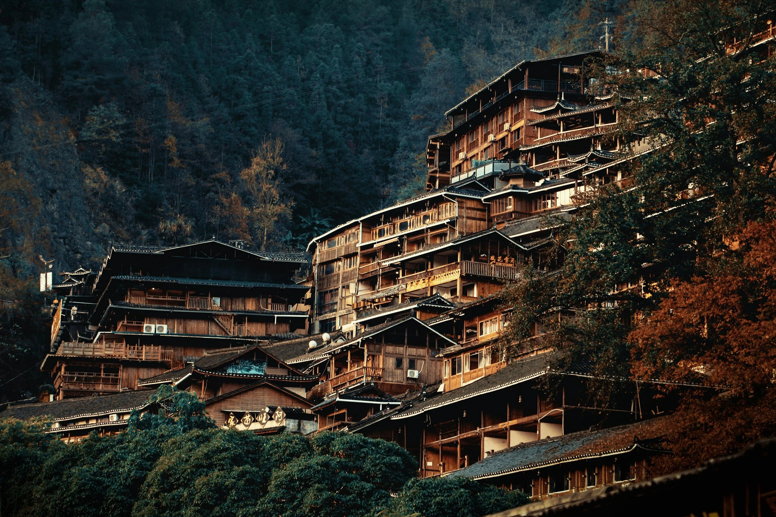 Multiple wooden houses built on a steep hillside surrounded by trees with fall foliage, set against a forested mountain background in Guizhou, China.