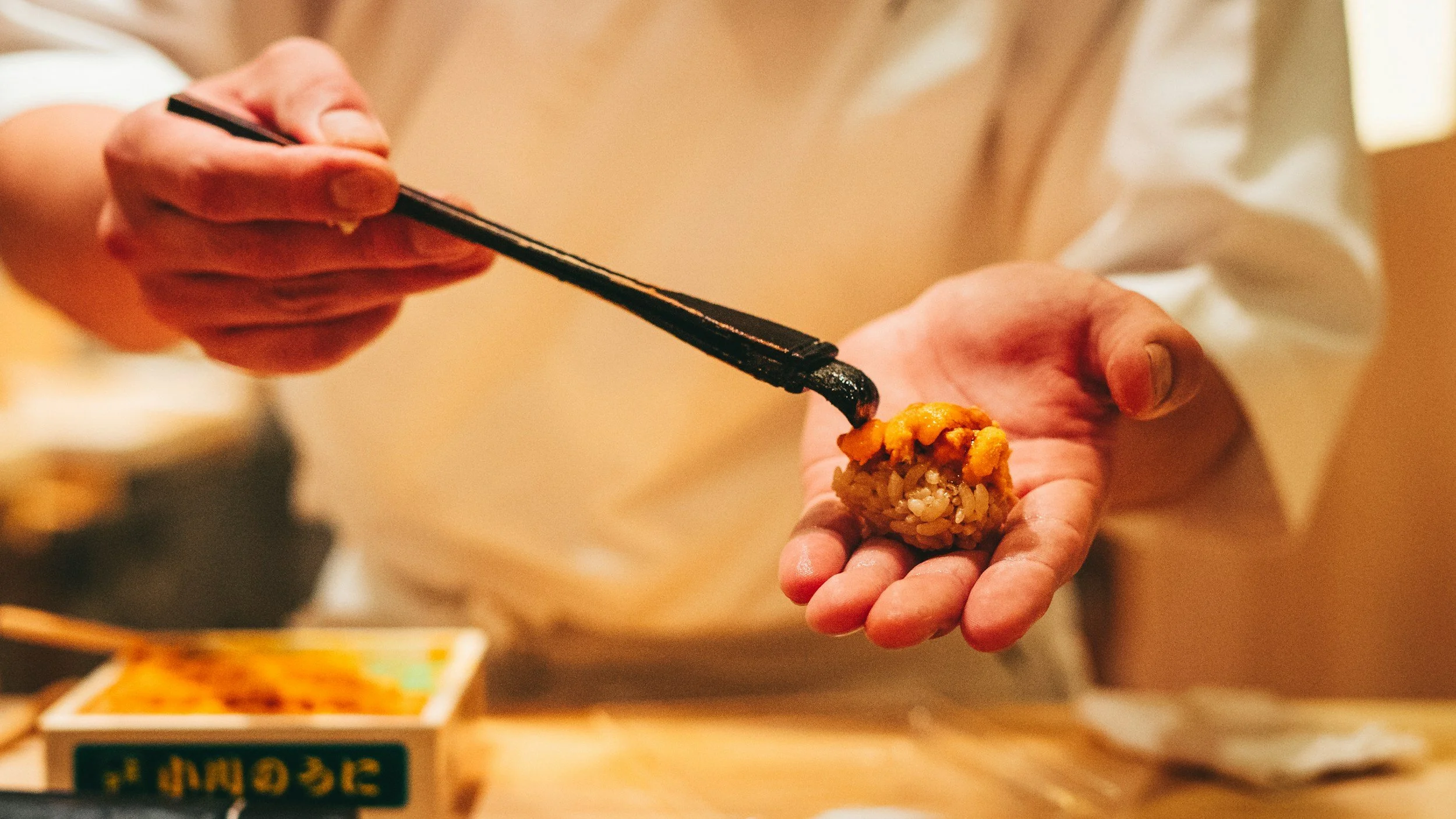 A person using chopsticks to place a piece of sear urchin uni sushi on their hand, with a sushi container and a wooden table in the background.