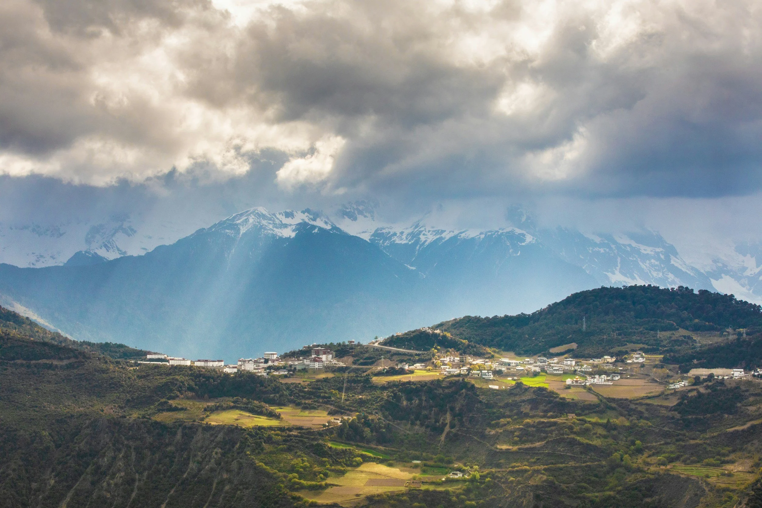 Landscape of mountains with snow-capped peaks under cloudy sky, with green hills and small white buildings in the foreground.