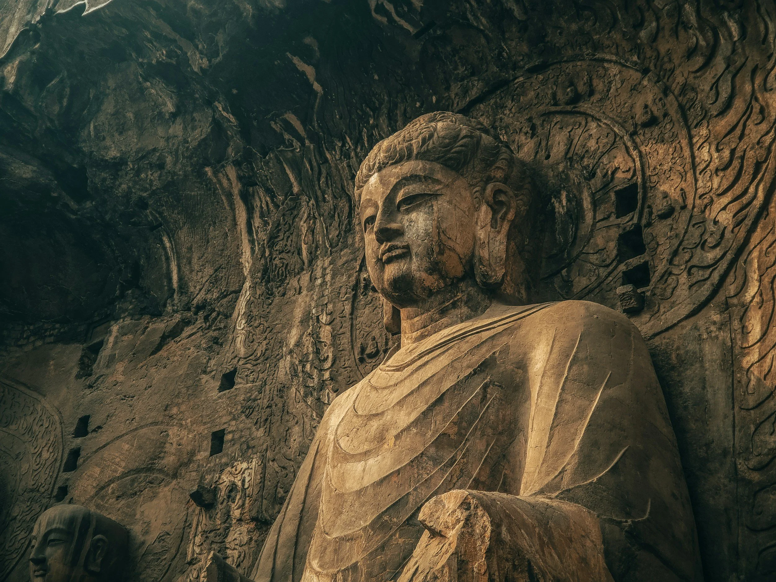 Ancient stone statue of a seated Buddha carved into a cave wall, with intricate carvings and patterns surrounding it at Longmen Grottoes in Henan, China.