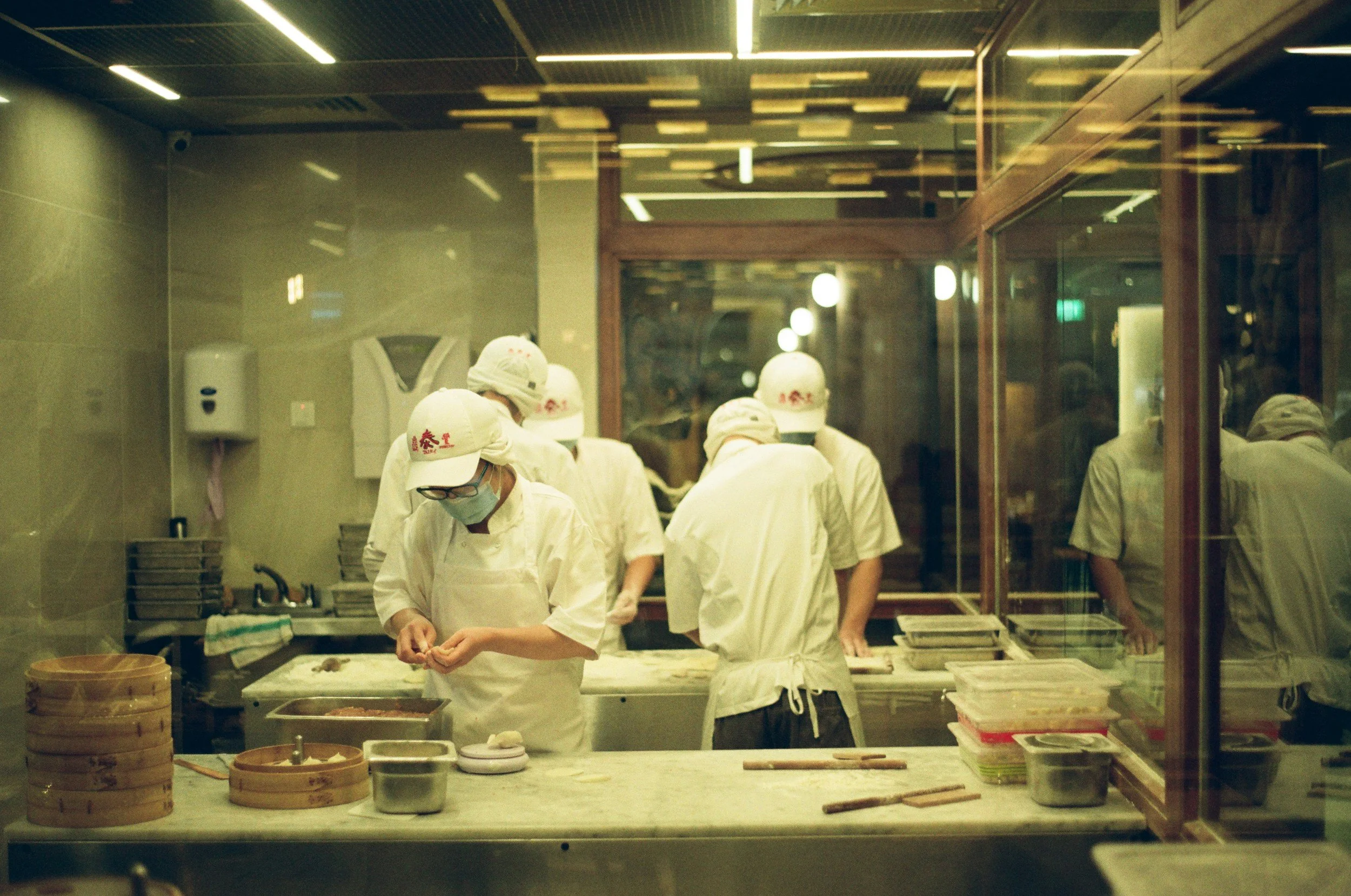 People working in a restaurant kitchen preparing xiaolongbao, wearing white uniforms, hats, and masks, seen through a glass wall.
